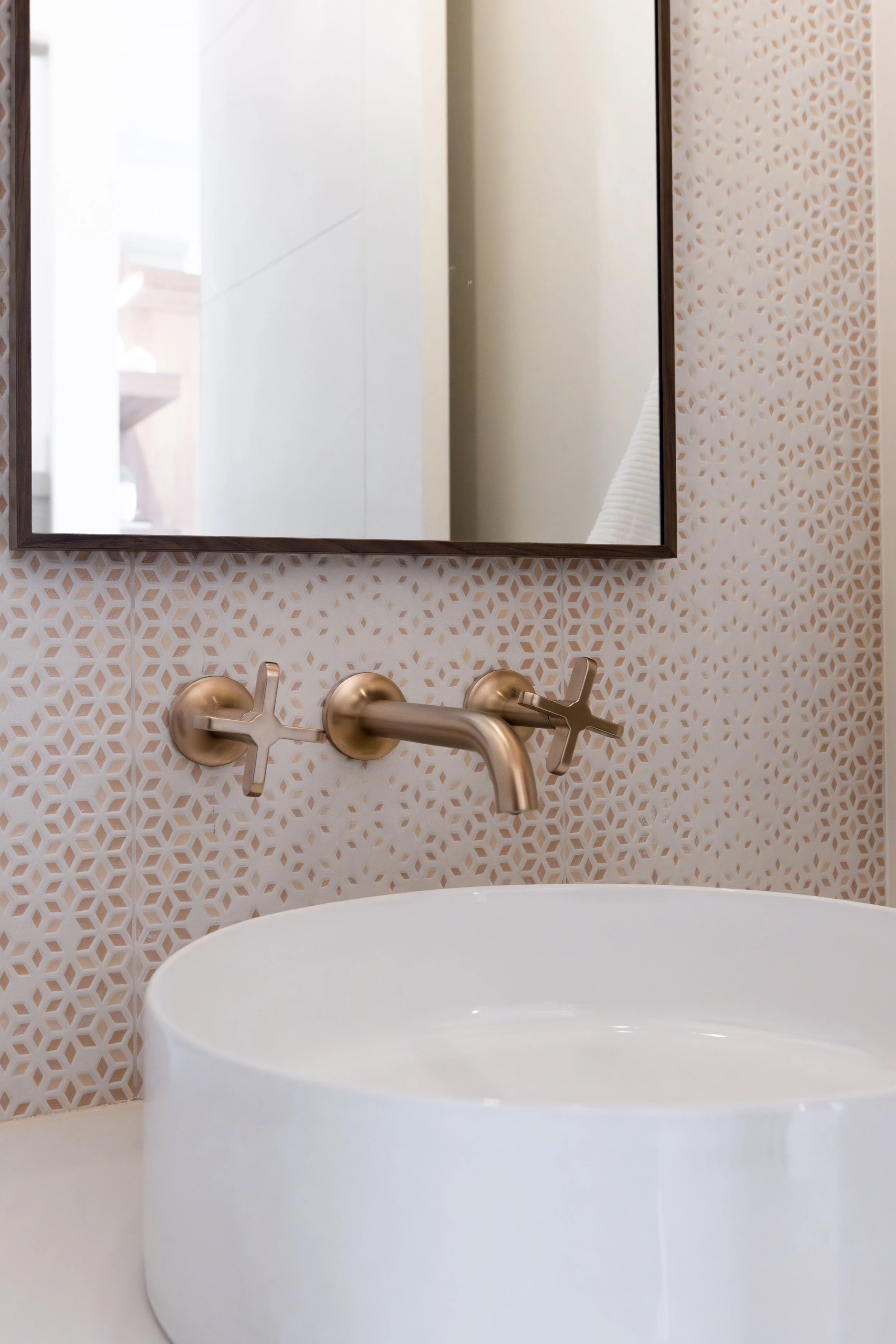 A bathroom sink with a rounded, white vessel basin mounted on a white countertop. Behind the sink, there's a wall with a beige and white geometric tile pattern. A rectangular mirror with a dark wooden frame hangs above the sink, reflecting a bright, 