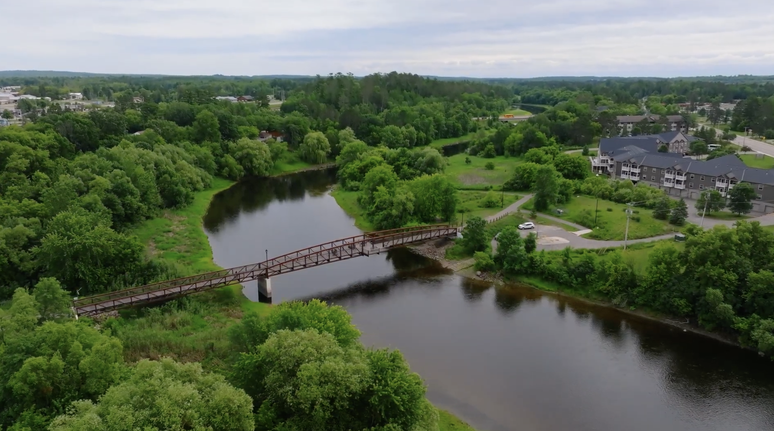 Aerial view of a river with a bridge, surrounded by dense green trees, residential buildings, and roads in a suburban area under a cloudy sky.