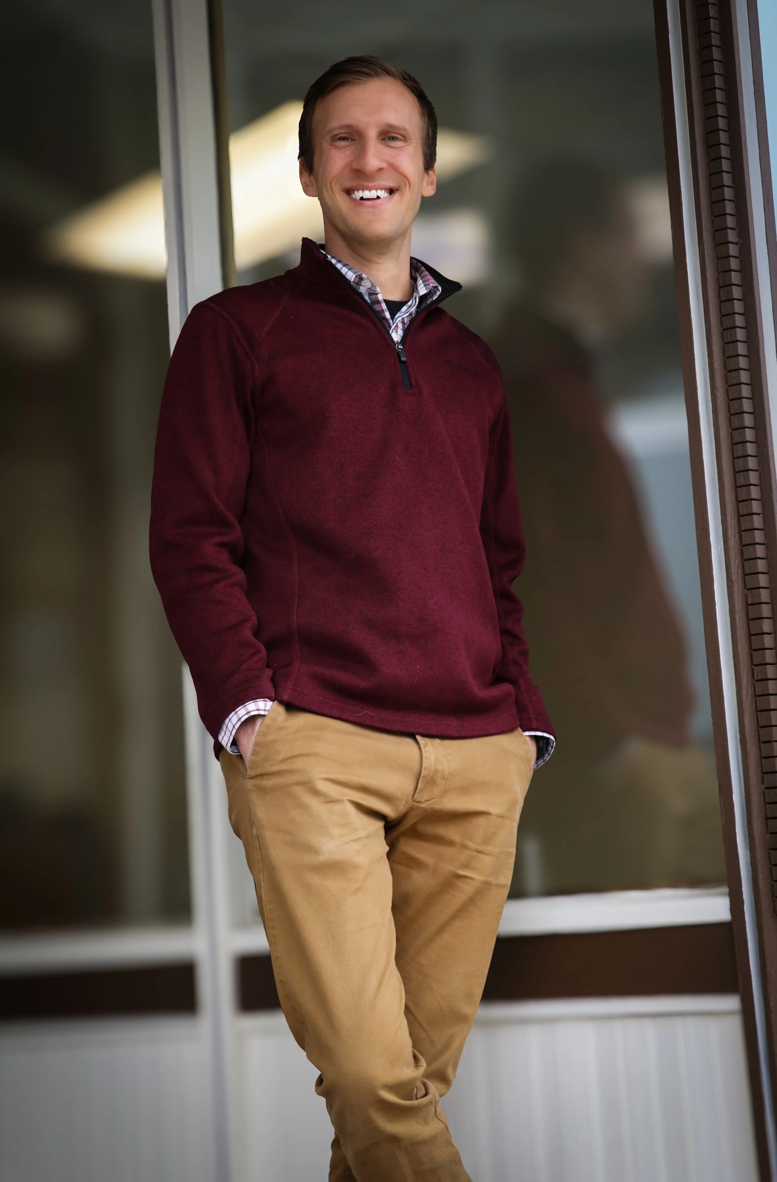 A smiling man wearing a burgundy sweater over a checkered shirt, standing with hands in pockets outside near a window.