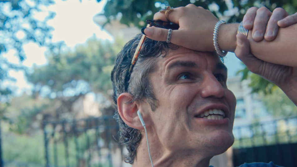 A man outdoors with headphones, sunglasses placed on his head, looking upward with a smile, and holding his hand to his forehead shielding his eyes from the sun.