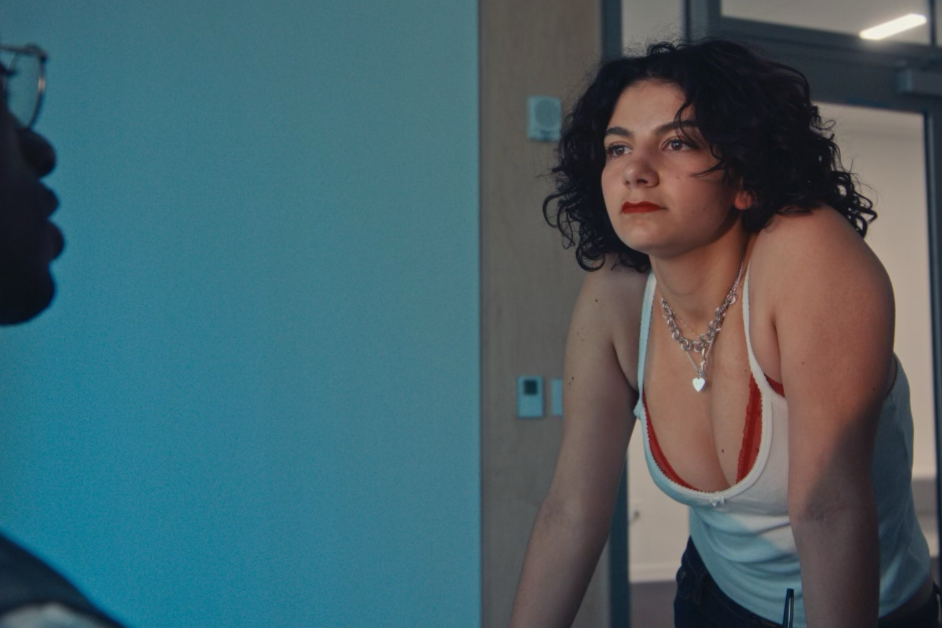 A woman with dark curly hair and red lipstick is leaning forward, looking intently at someone or something off-camera. She is wearing a white tank top with red accents and layered necklaces. The background shows part of a room with wooden walls and a