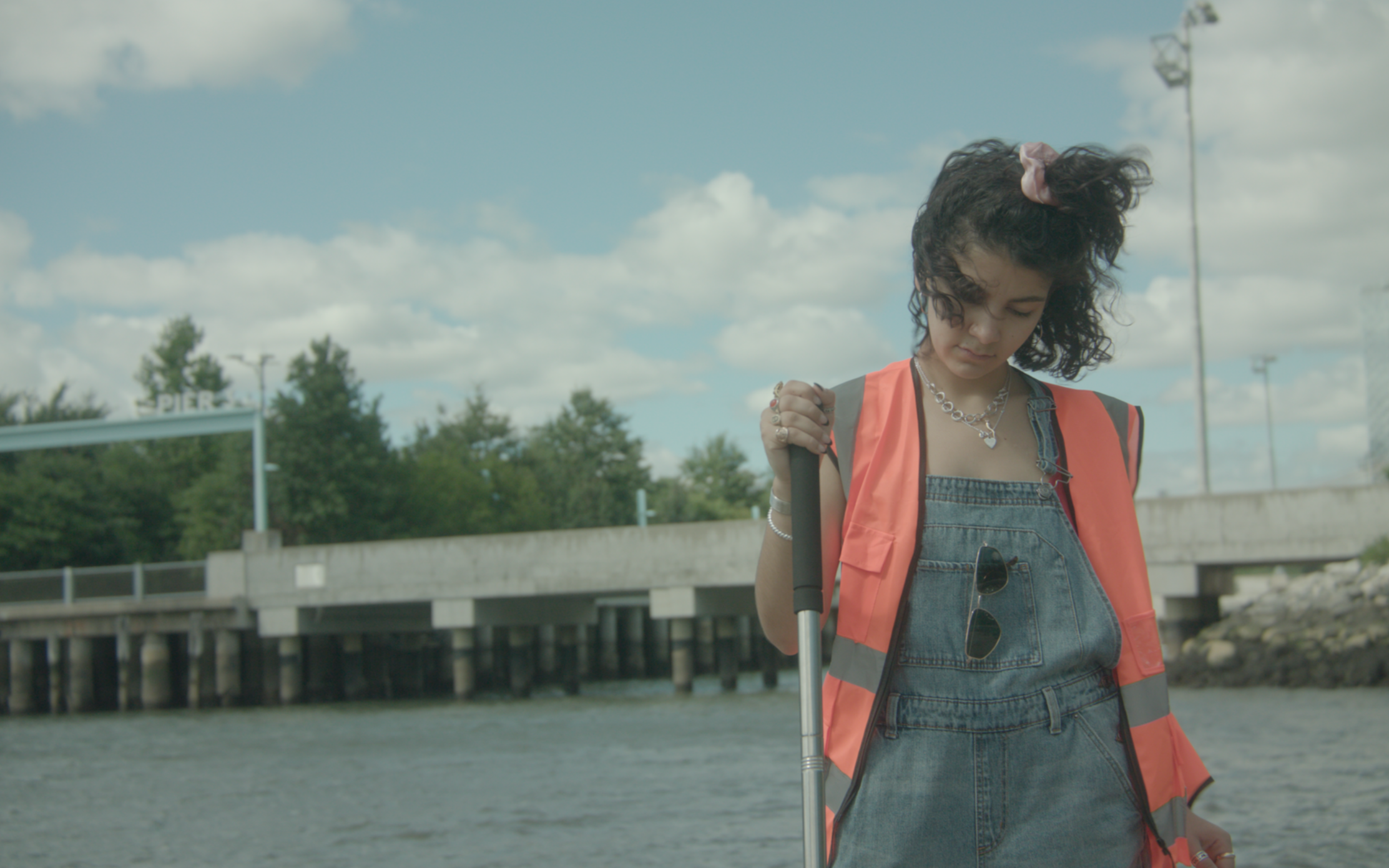 A woman with short curly hair wearing a pink vest over denim overalls and holding a cane, standing outdoors near a canal or river with a bridge and trees in the background, under a partly cloudy sky.