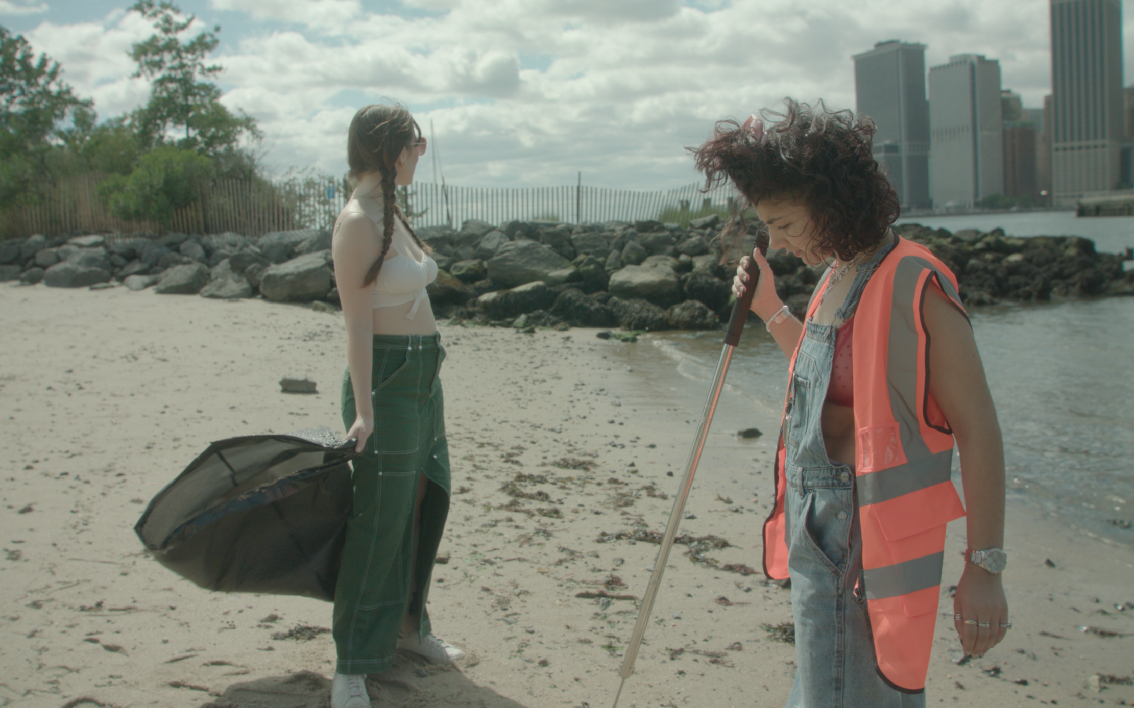 Two women on a beach, one with long brown hair and another with curly hair, digging in the sand with a metal stick, with city buildings and cloudy sky in the background.