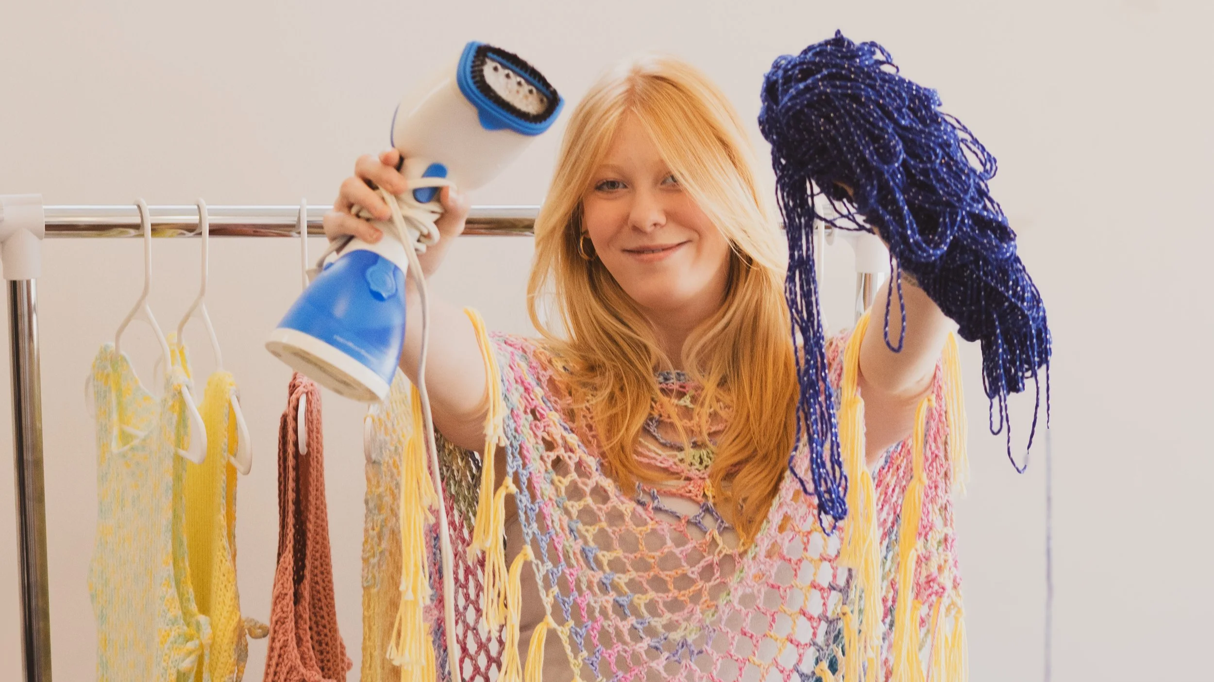 Maggie Gaut holding a steamer and a bundle of blue yarn in front of a rack of crochet ponchos