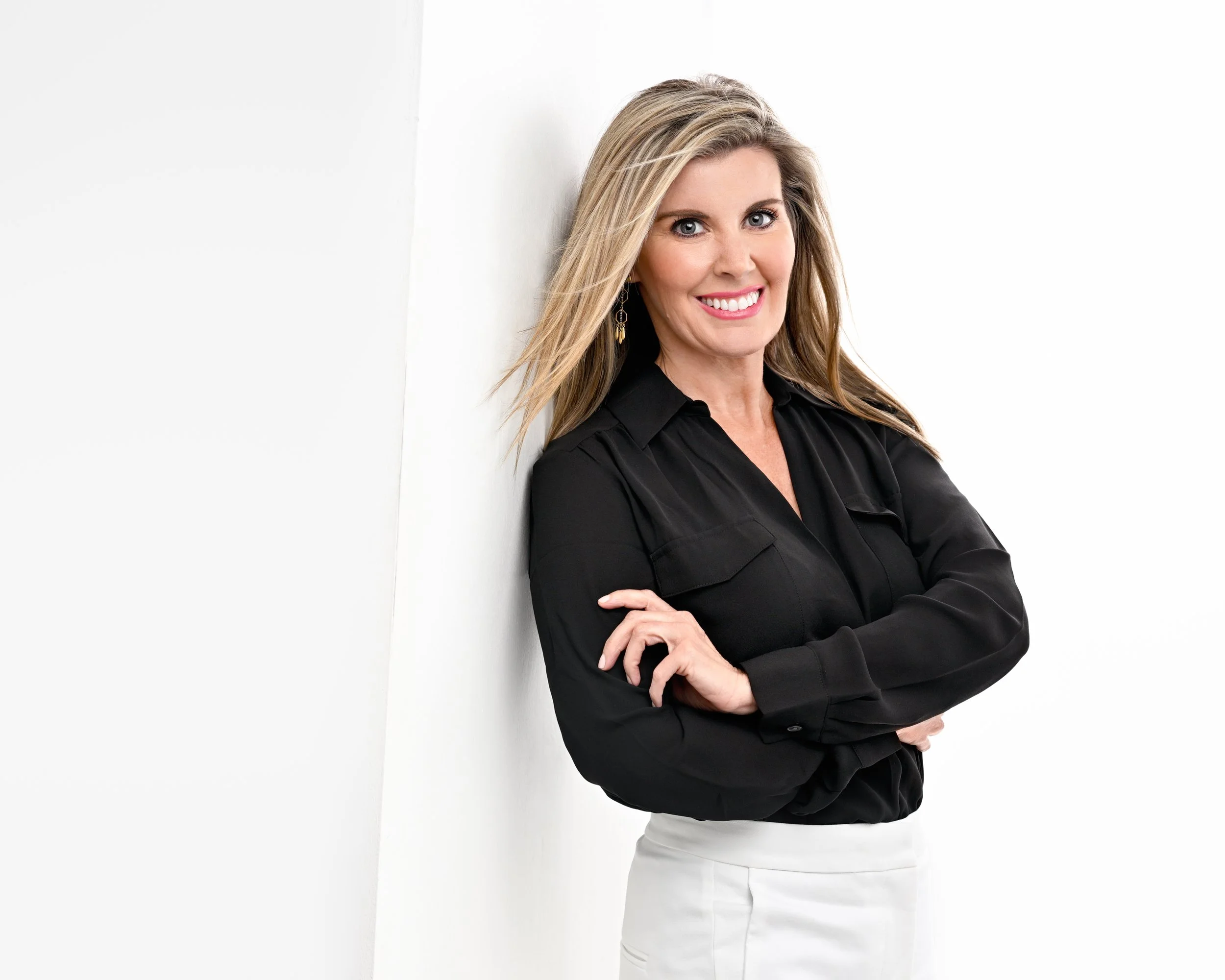 A woman with blonde hair smiling, wearing a black blouse and white pants, leaning against a white wall.