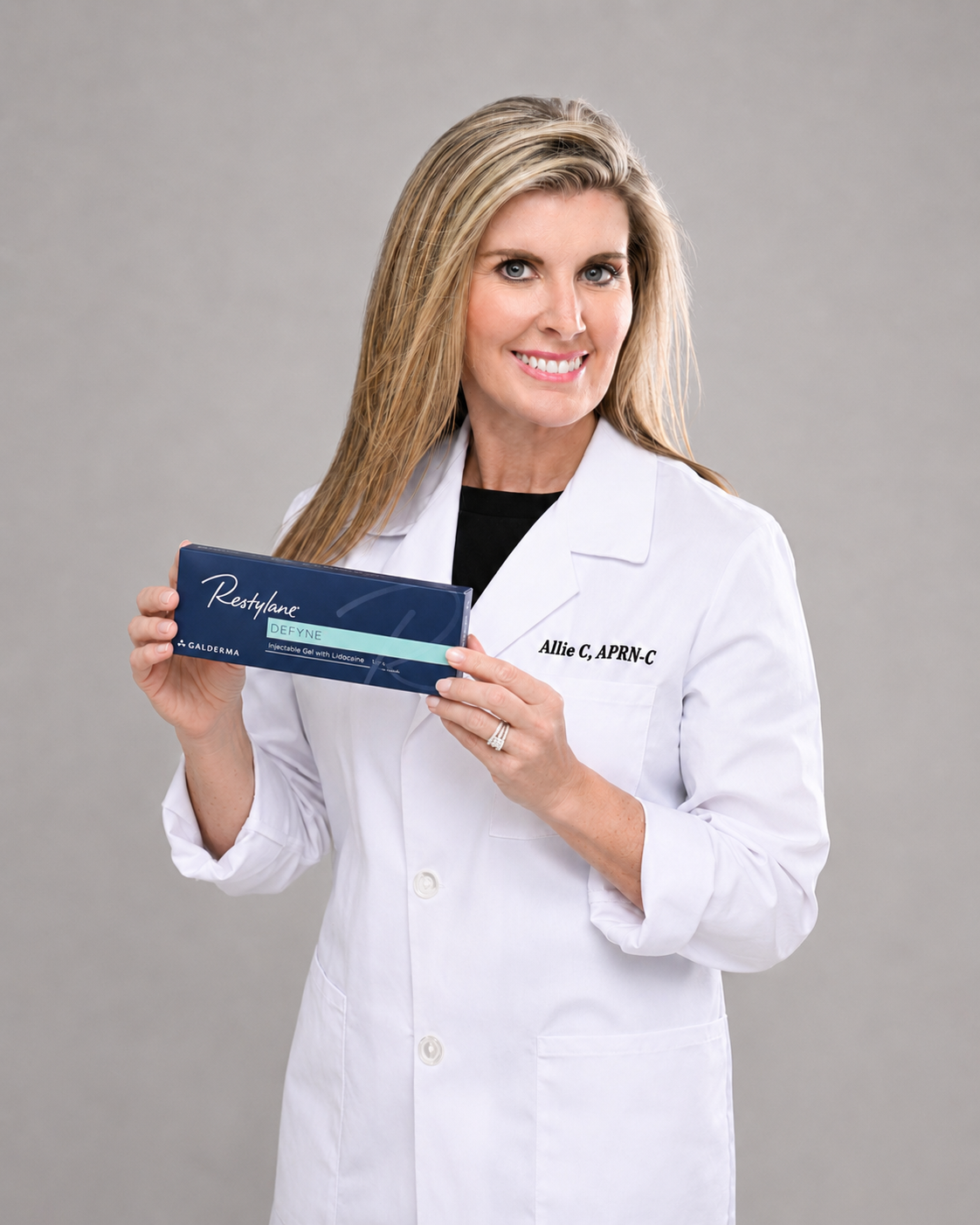A woman in a white medical coat holding a box of Restylane Defyne dermal filler, smiling at the camera against a grey background.