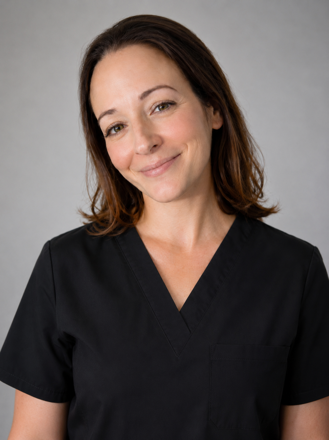 A woman with shoulder-length brown hair smiling, wearing a black medical scrub top, against a plain gray background.