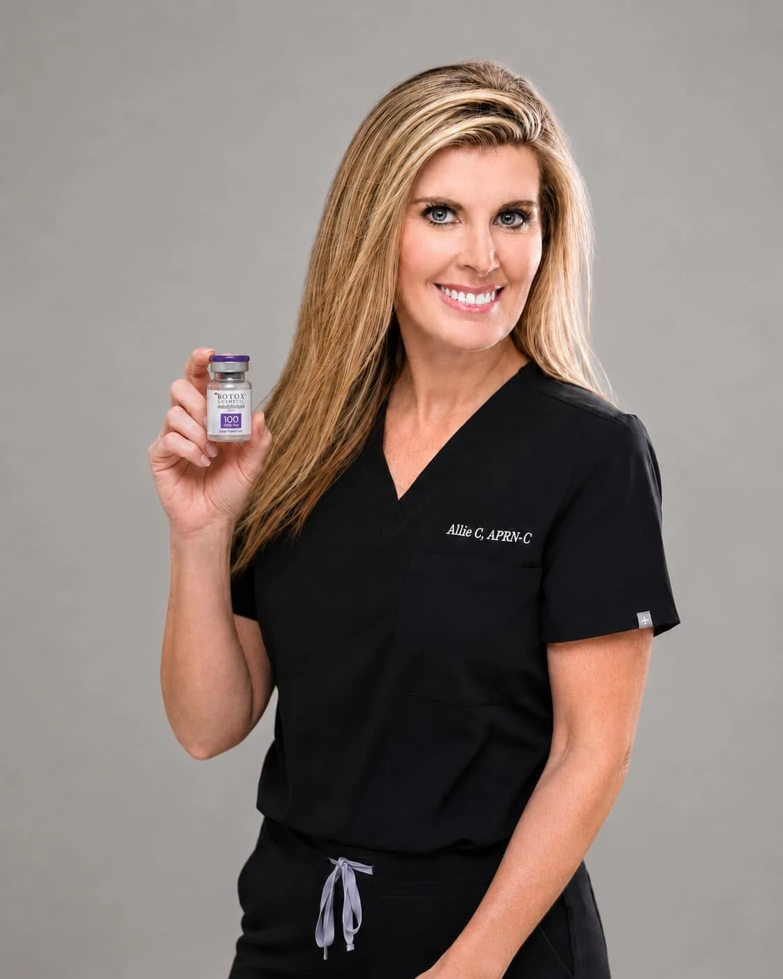 A woman in medical scrubs holding a small vial labeled 'Botox' with a purple cap, smiling at the camera against a plain gray background.