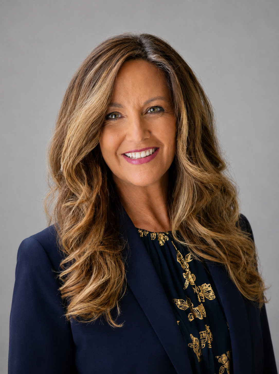 A woman with long, wavy, light brown hair smiling at the camera. She wears a dark blazer over a blouse with a gold floral pattern, against a plain gray background.