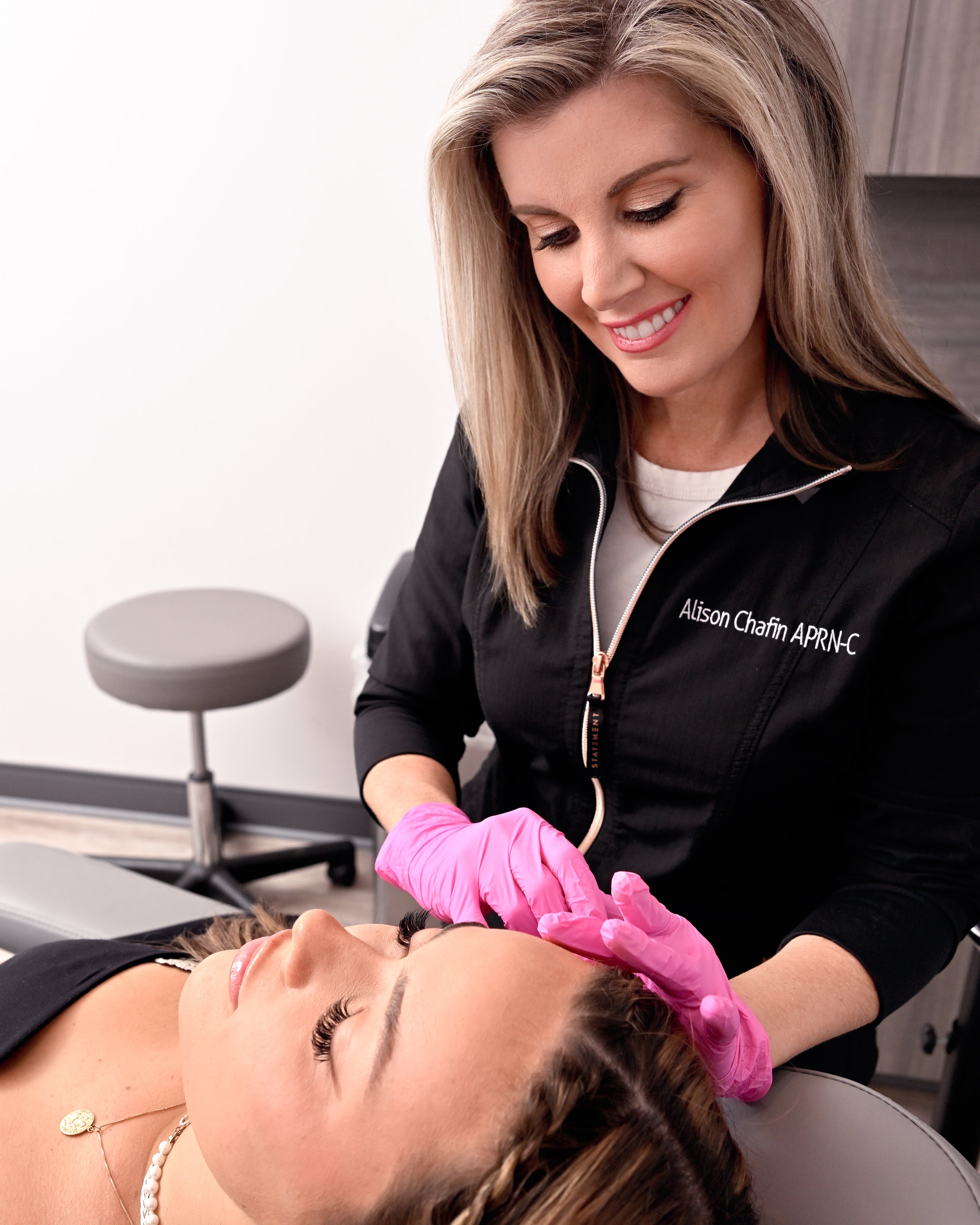 A woman receiving a cosmetic procedure while lying on a treatment bed, with a healthcare professional wearing pink gloves and a black jacket, smiling.