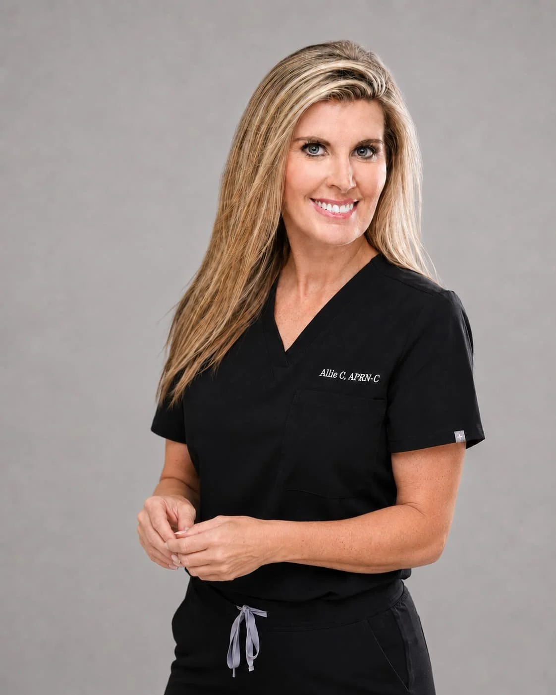 A woman with long blonde hair, smiling, wearing a black medical scrub top with the name "Allie C, APRN-C" embroidered on it, standing in front of a plain gray background.