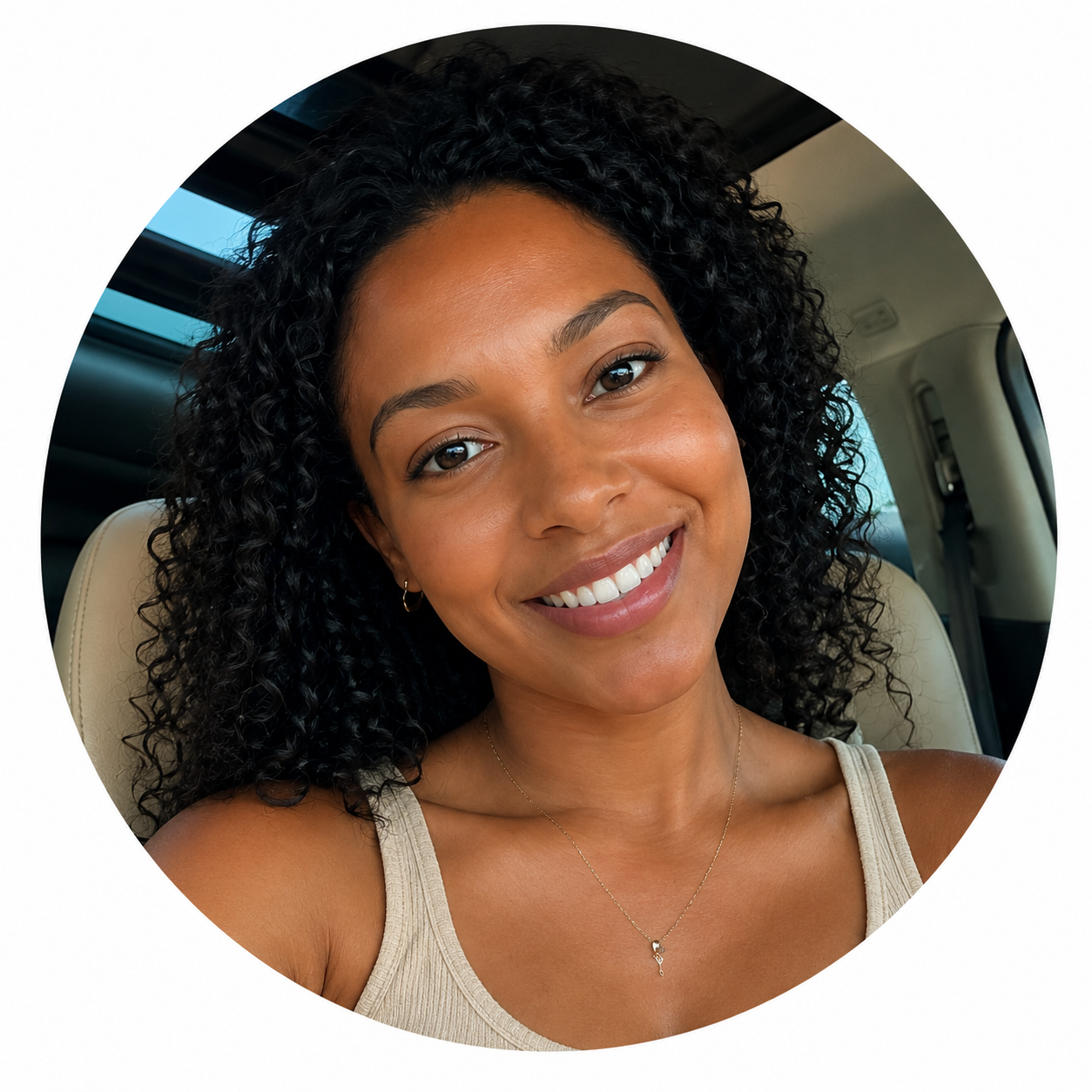 A woman with dark curly hair smiling, sitting inside a vehicle, wearing a beige tank top and a delicate necklace.