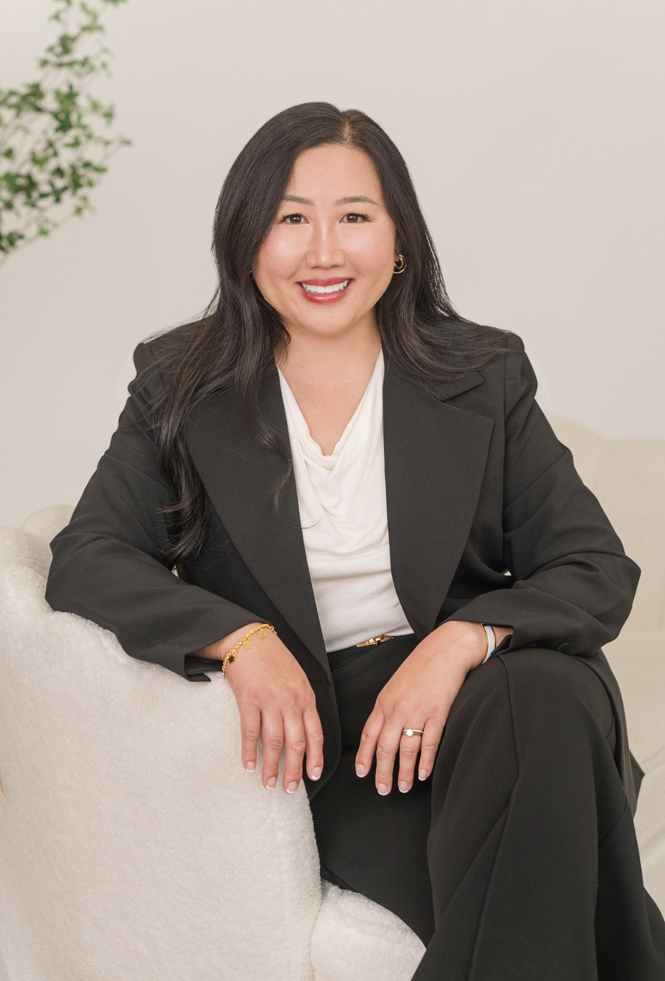 A woman with long black hair, smiling, wearing a black blazer and white top, sitting on a beige sofa with a white background and a green leafy plant in the background.