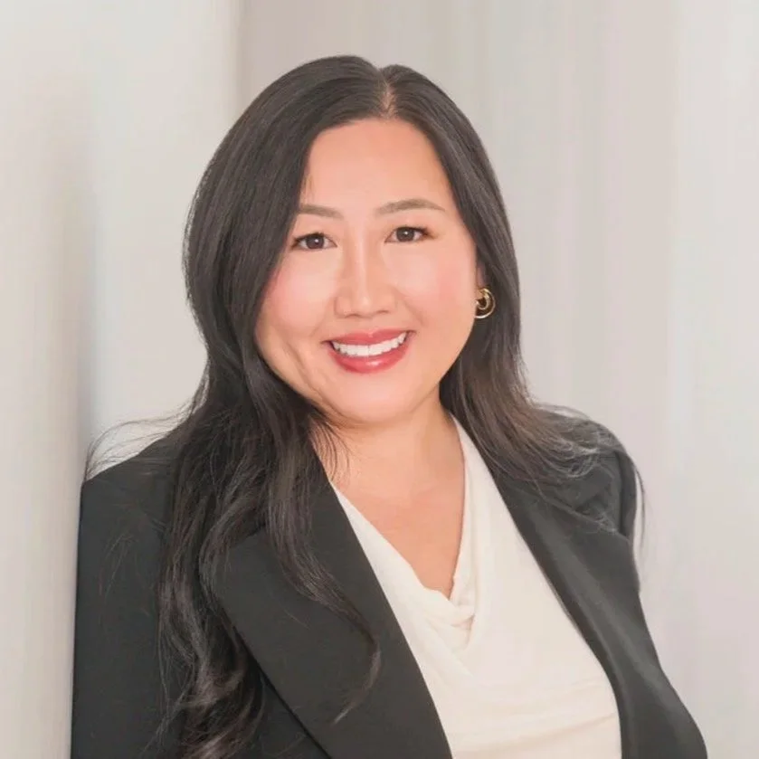 Portrait of a woman with long dark hair, smiling, wearing a black blazer over a white top, standing against a light background.