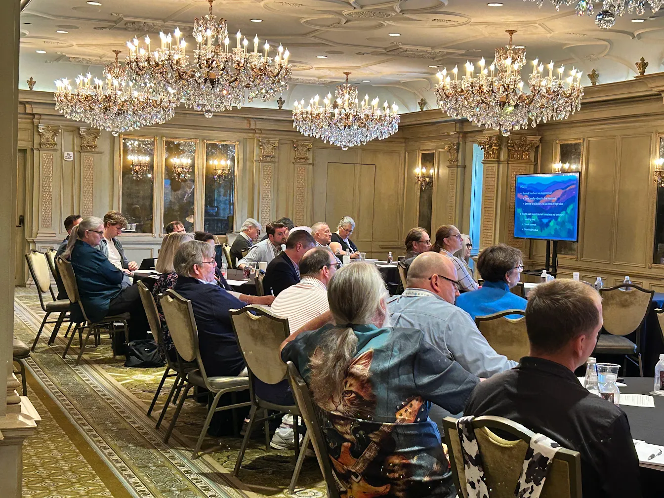 Attendees gather for a presentation in an ornate ballroom with crystal chandeliers and a presentation screen.