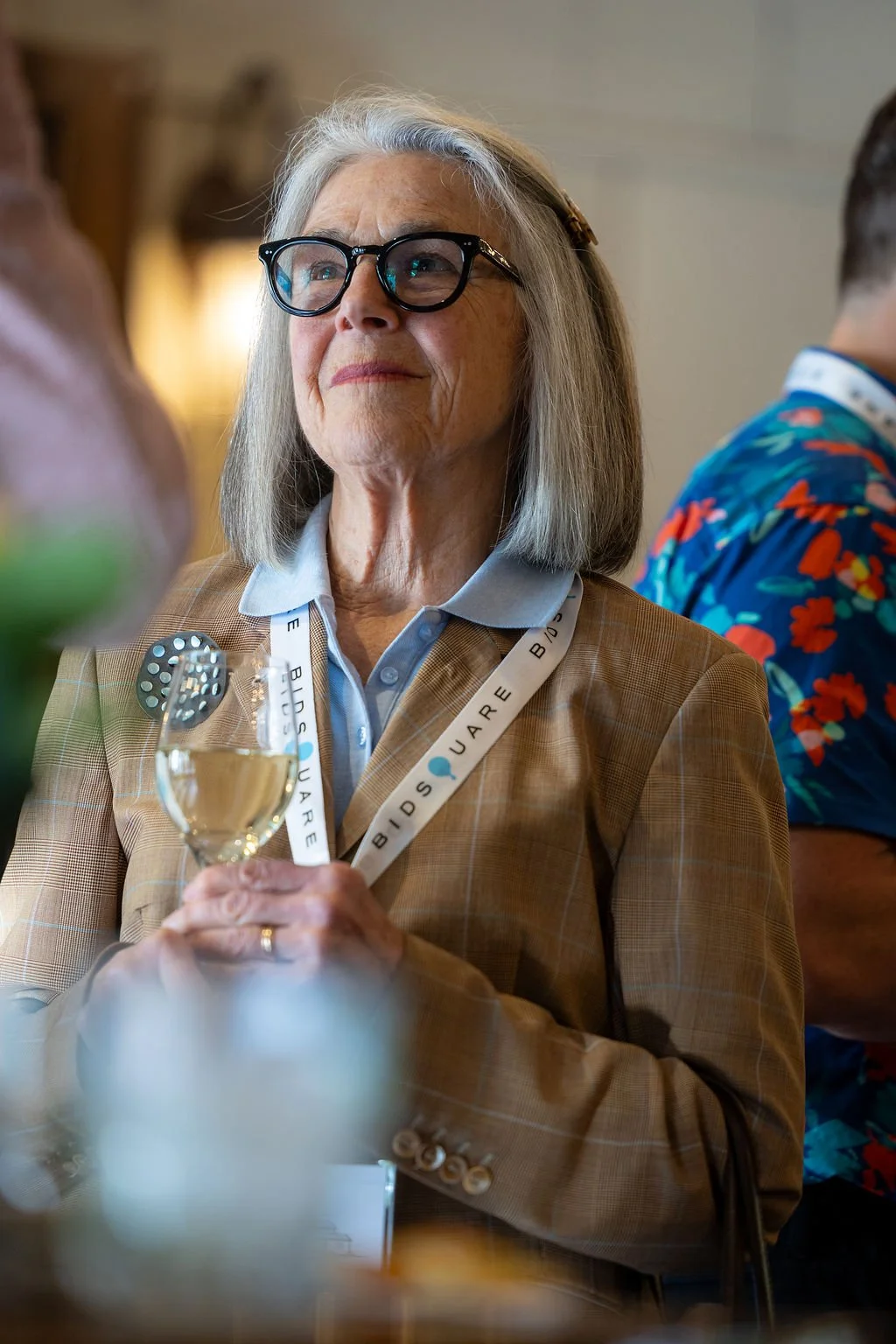 Gray-haired woman wearing glasses and a plaid blazer holds a glass of white wine at an indoor event.