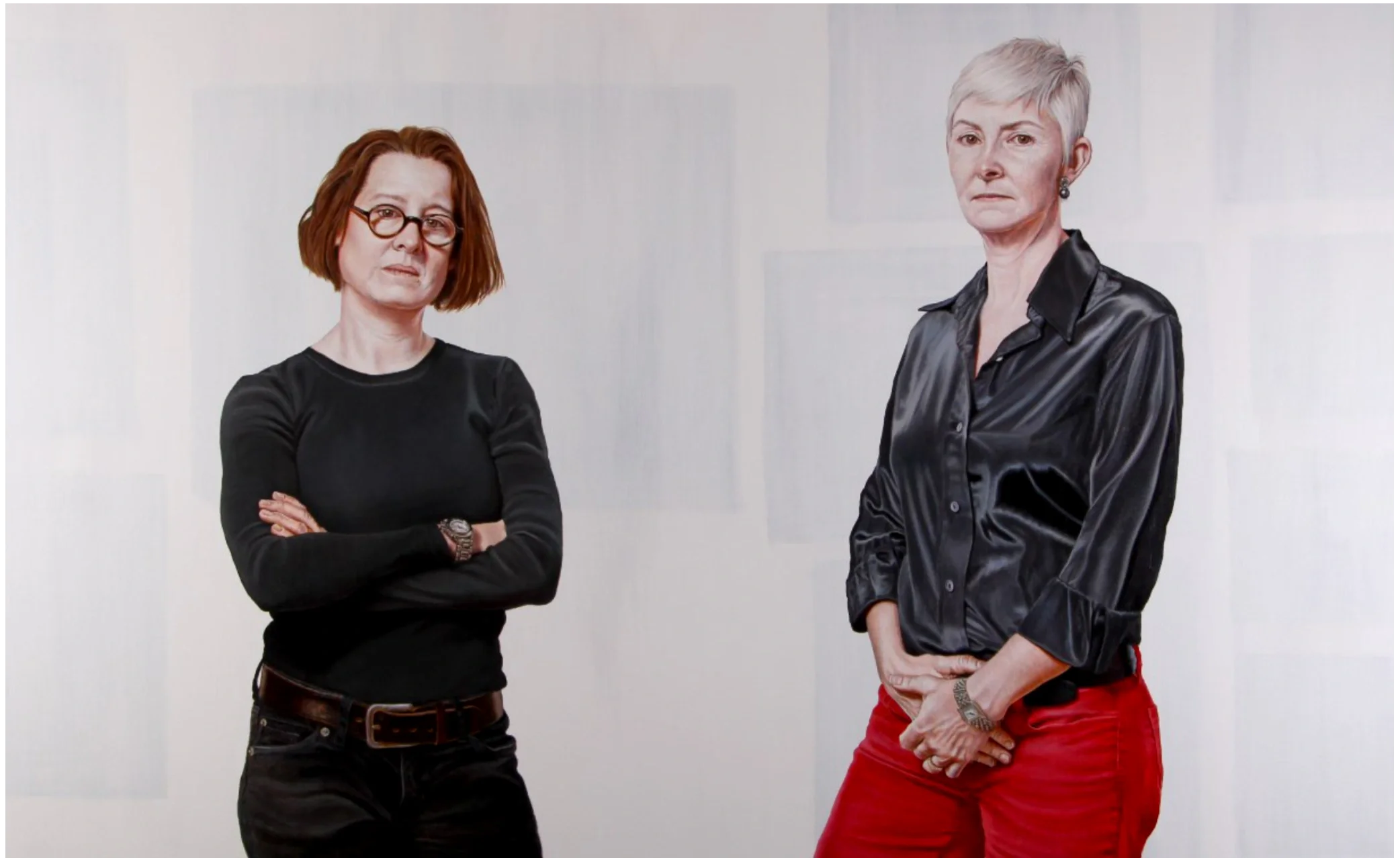 Portrait of two women standing against a light background, one wearing glasses and a black outfit, the other wearing a black shirt and red pants