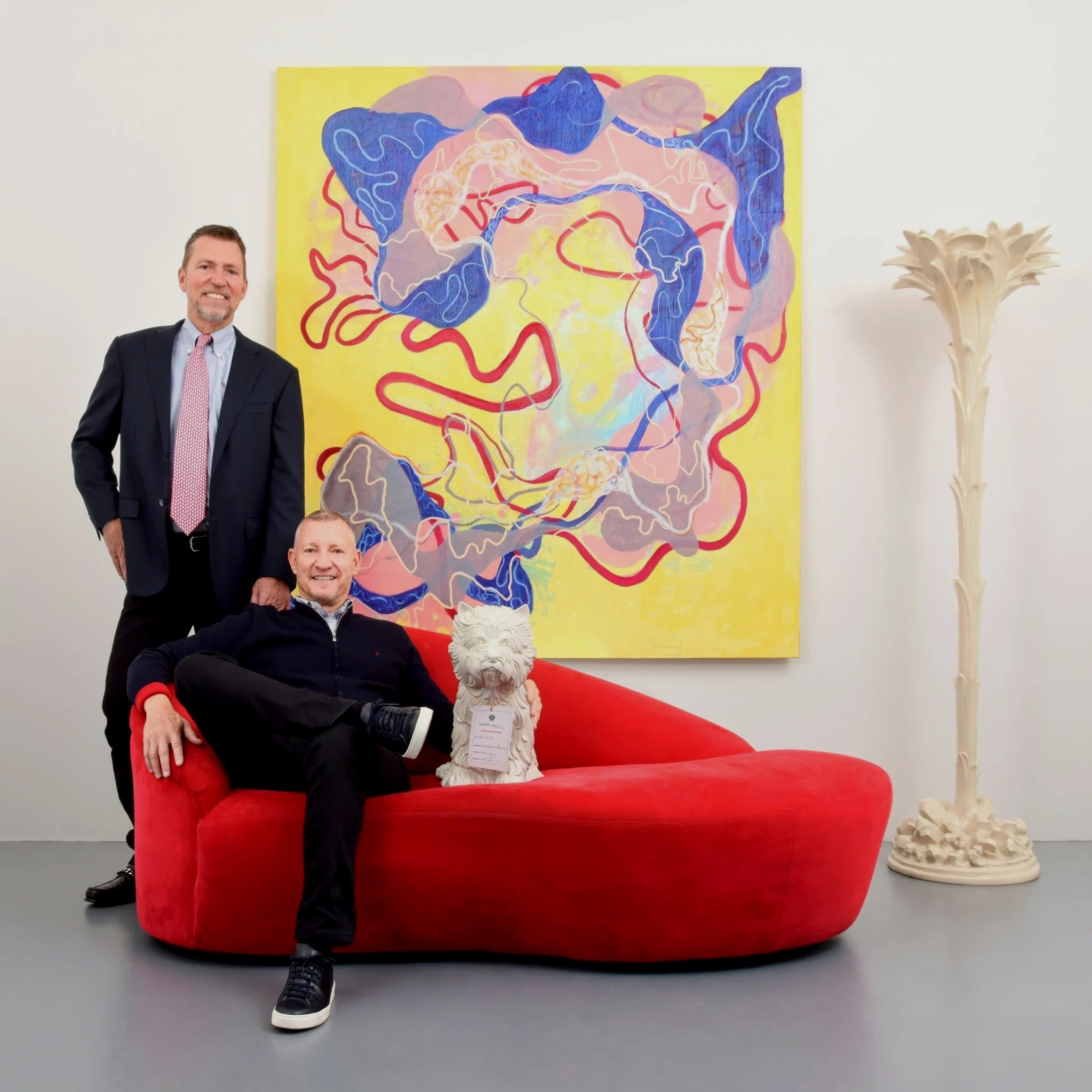 Two men pose in a modern room with a large abstract painting, red sofa, white dog statue, and sculptural floor lamp.