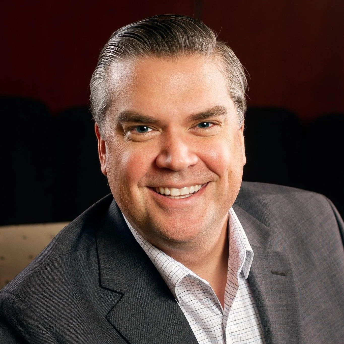 A smiling man in a gray suit and white checkered shirt, sitting against a dark background.