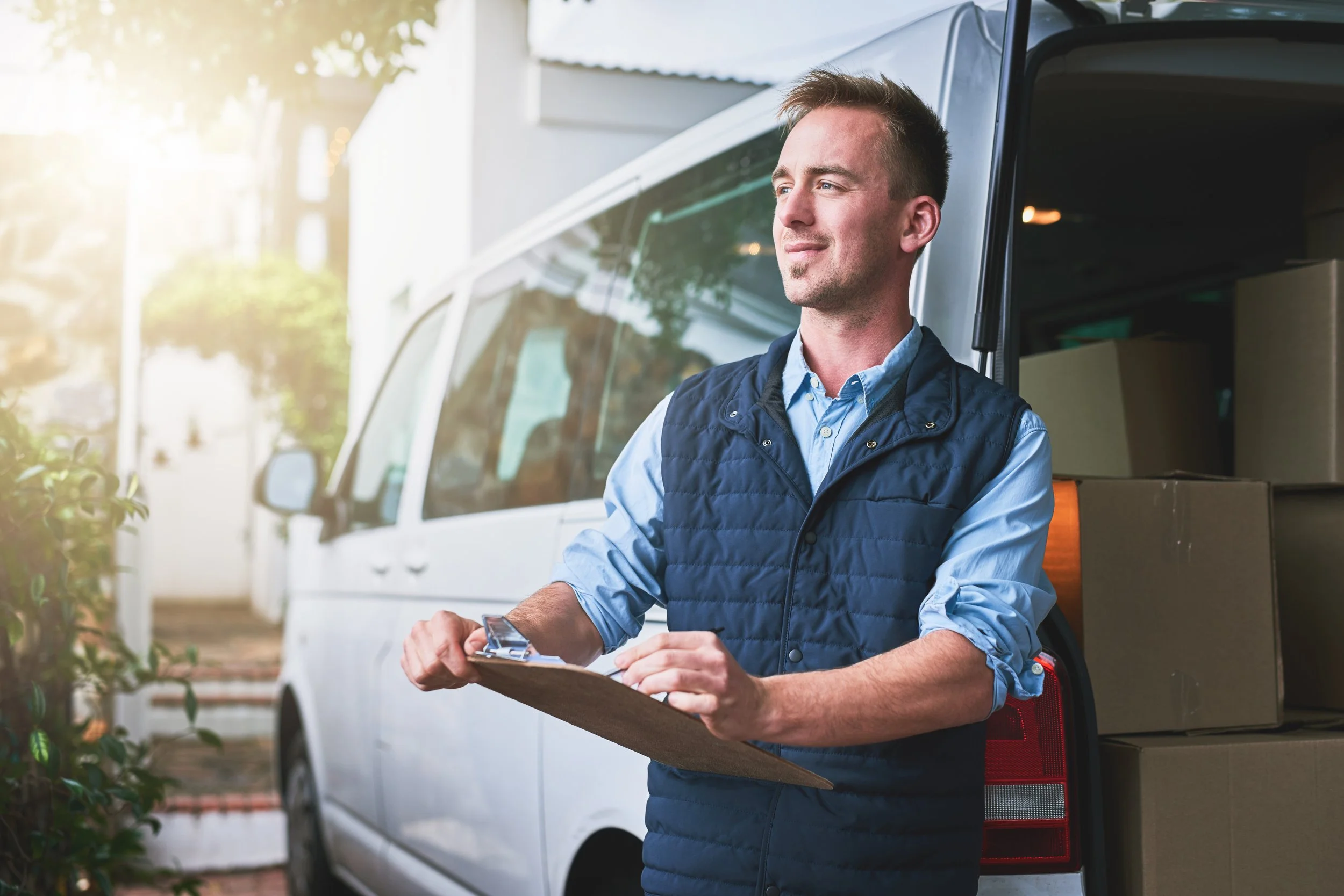 A man in a blue vest and blue shirt holding a clipboard, leaning against the back of a white delivery van, looking off to the side with a slight smile, in a suburban area with trees and a building in the background.