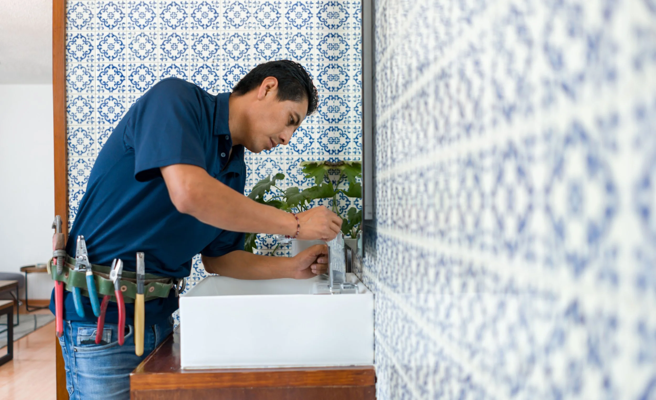 A man wearing a blue shirt and jeans, equipped with tools hanging from his waist, is fixing a wall-mounted fixture or appliance in a room with blue and white patterned tiles. He is focused on his task, leaning over the sink area, with greenery visible in the background.