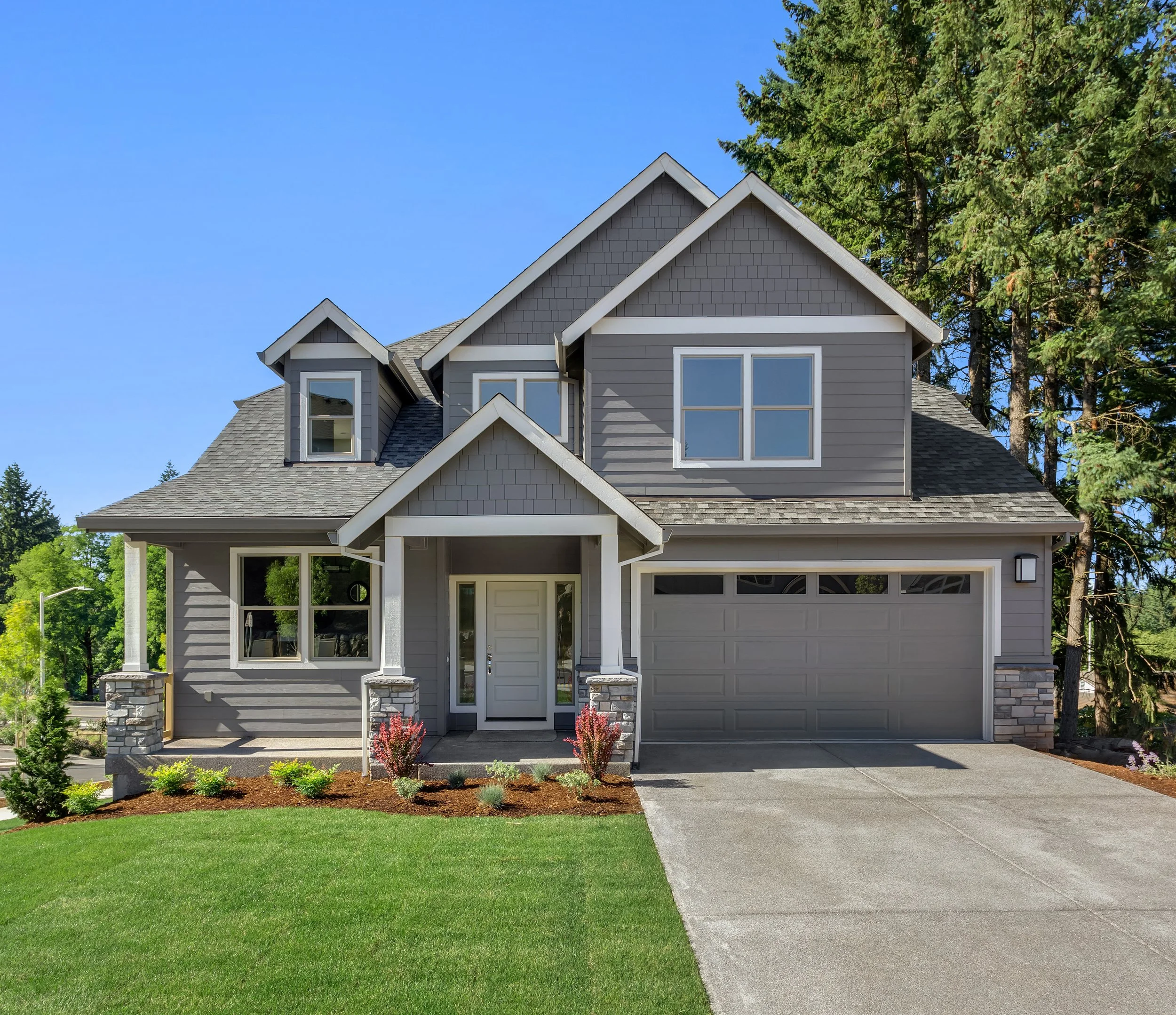 A modern gray house with large windows, a front porch, and a well-maintained lawn, surrounded by trees under a clear blue sky.