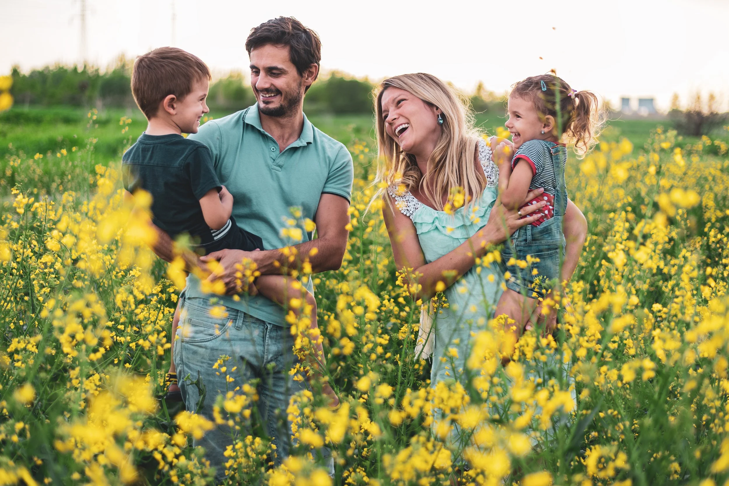 Family of four, including two children, enjoying time together in a field of yellow flowers during sunset.