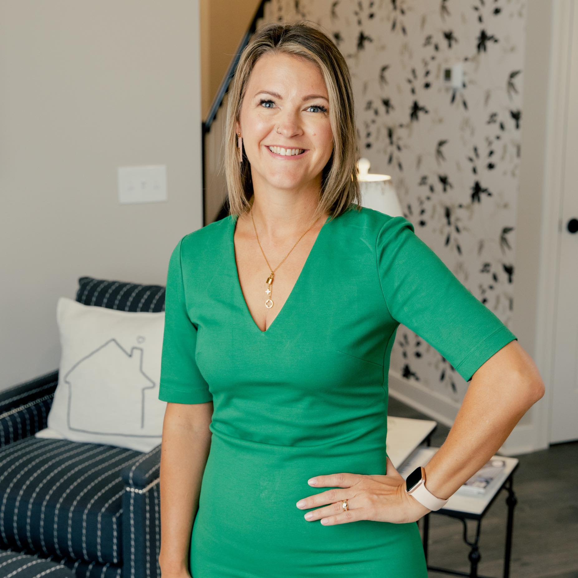 A smiling woman with shoulder-length blonde hair, wearing a green V-neck dress, gold necklaces, and jewelry, standing in a modern living room with black and white patterned wallpaper, a black and white sofa, and decorative pillows.