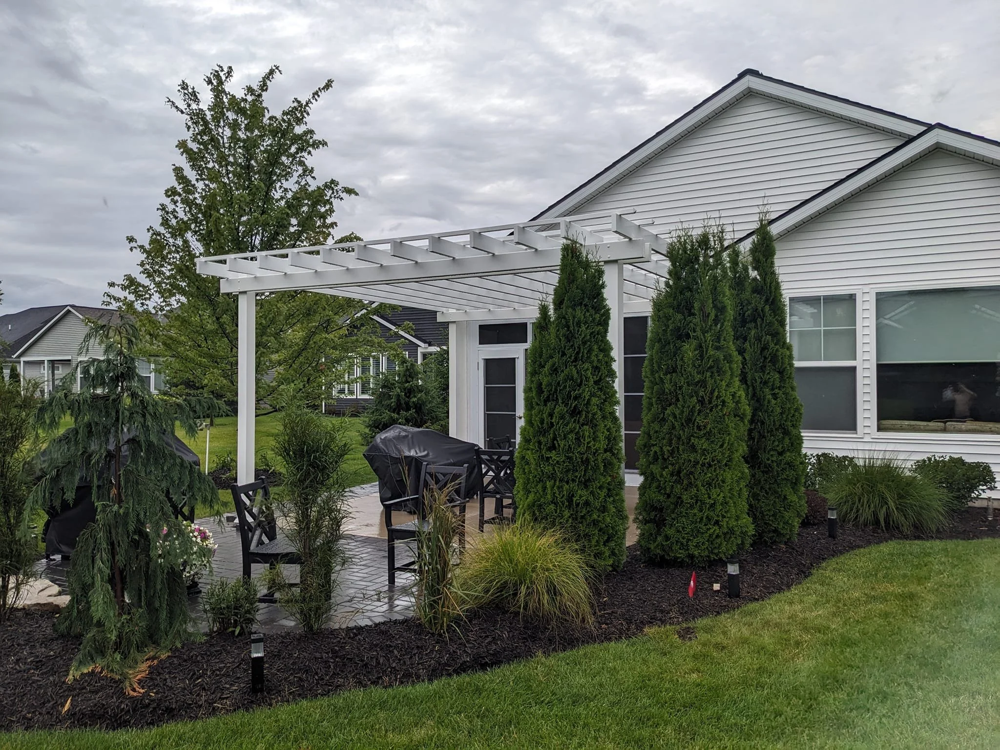 A backyard patio with a black table and multiple black chairs, surrounded by green shrubs and plants, with a white house in the background and a pergola overhead. There are trees and neighboring houses visible under a cloudy sky.
