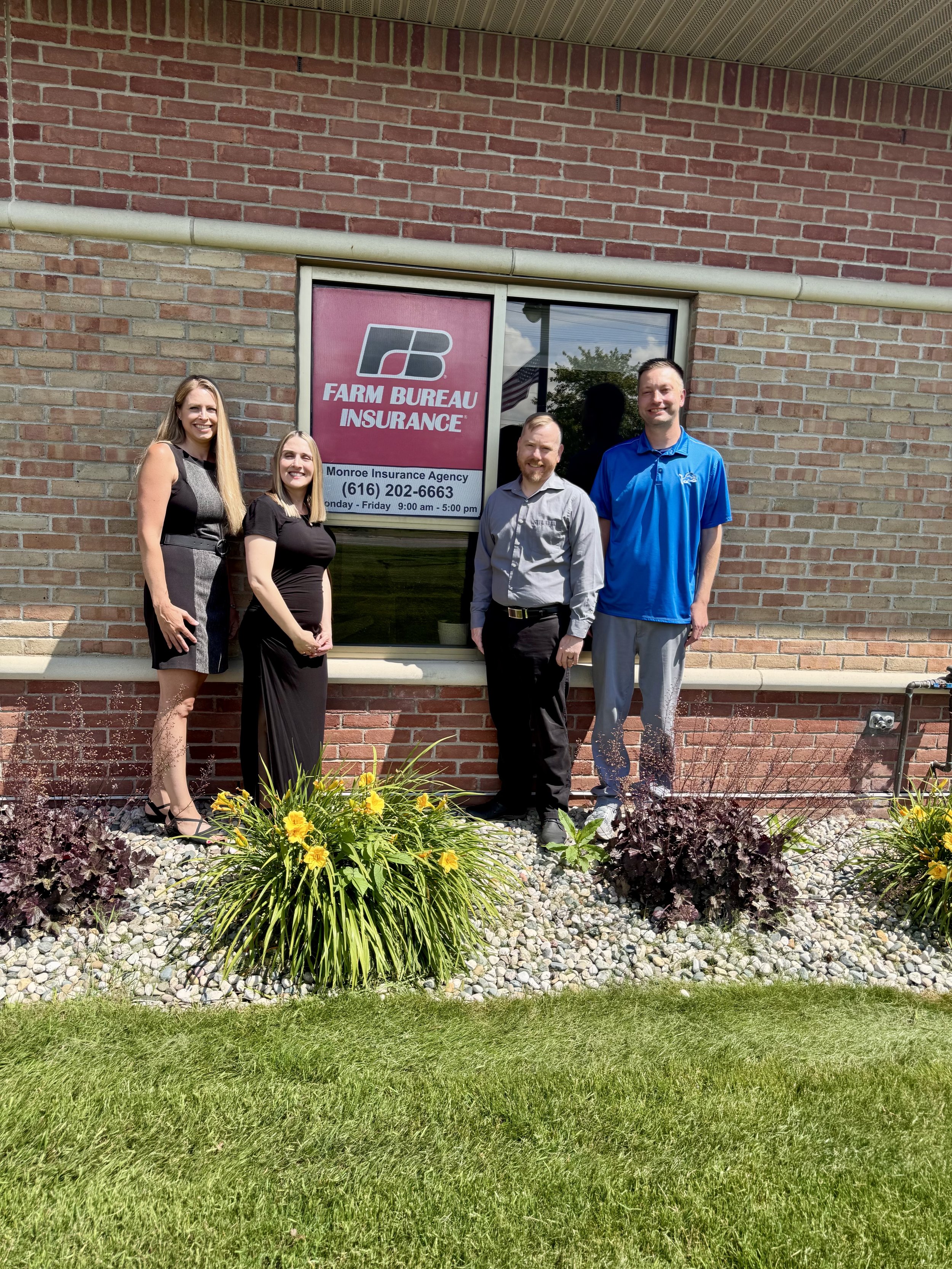 Four people standing in front of a brick building with a window that has a sign reading "Farm Bureau Insurance" and a phone number. The group includes two women and two men, all smiling. There are yellow flowers and decorative plants in the foreground.