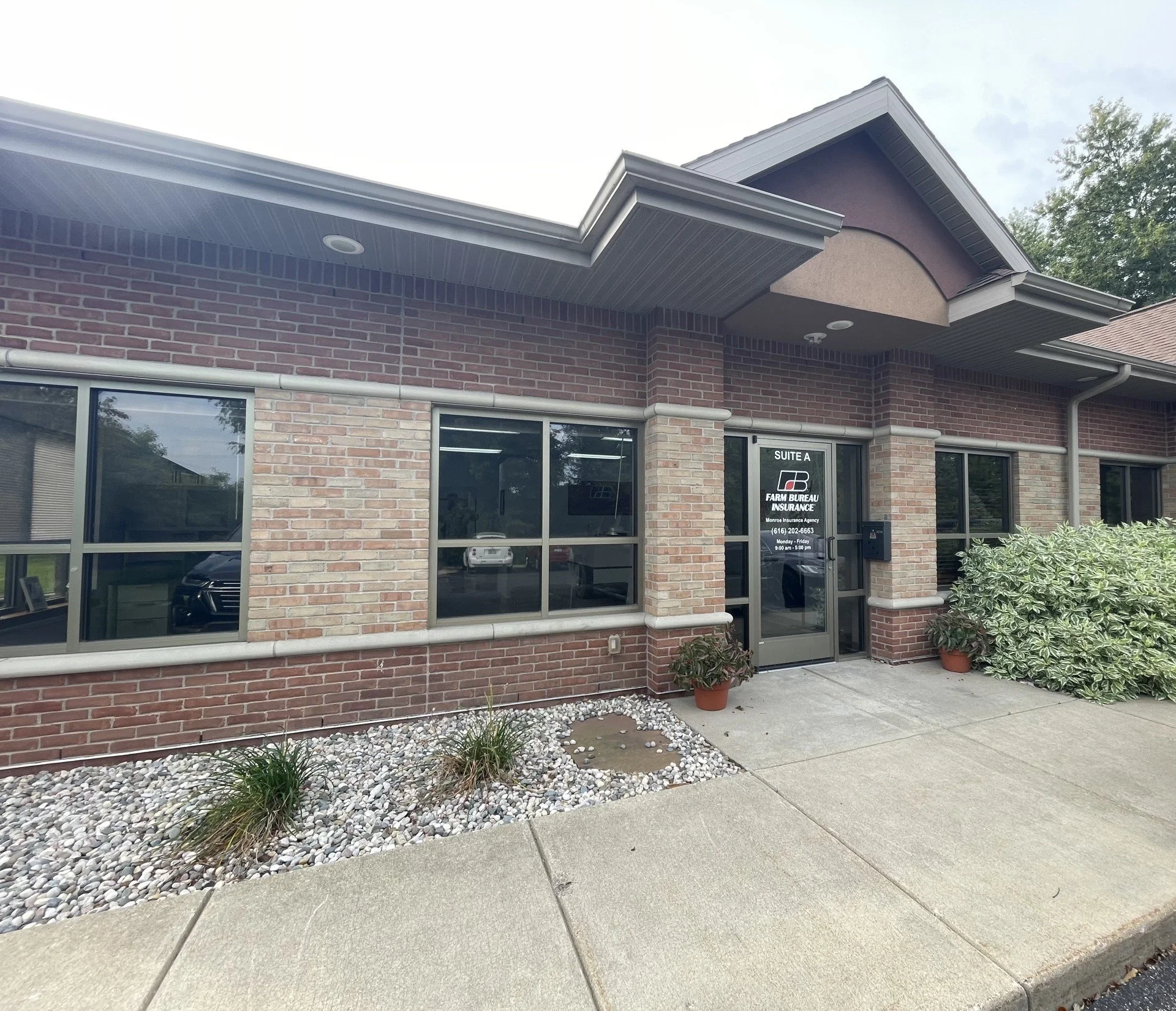 Front exterior of a brick commercial building with a sign on the door that reads 'SUITE A FARM BUREAU INSURANCE.' Potted plants and small landscaping rocks are near the entrance, with a sidewalk in the foreground.