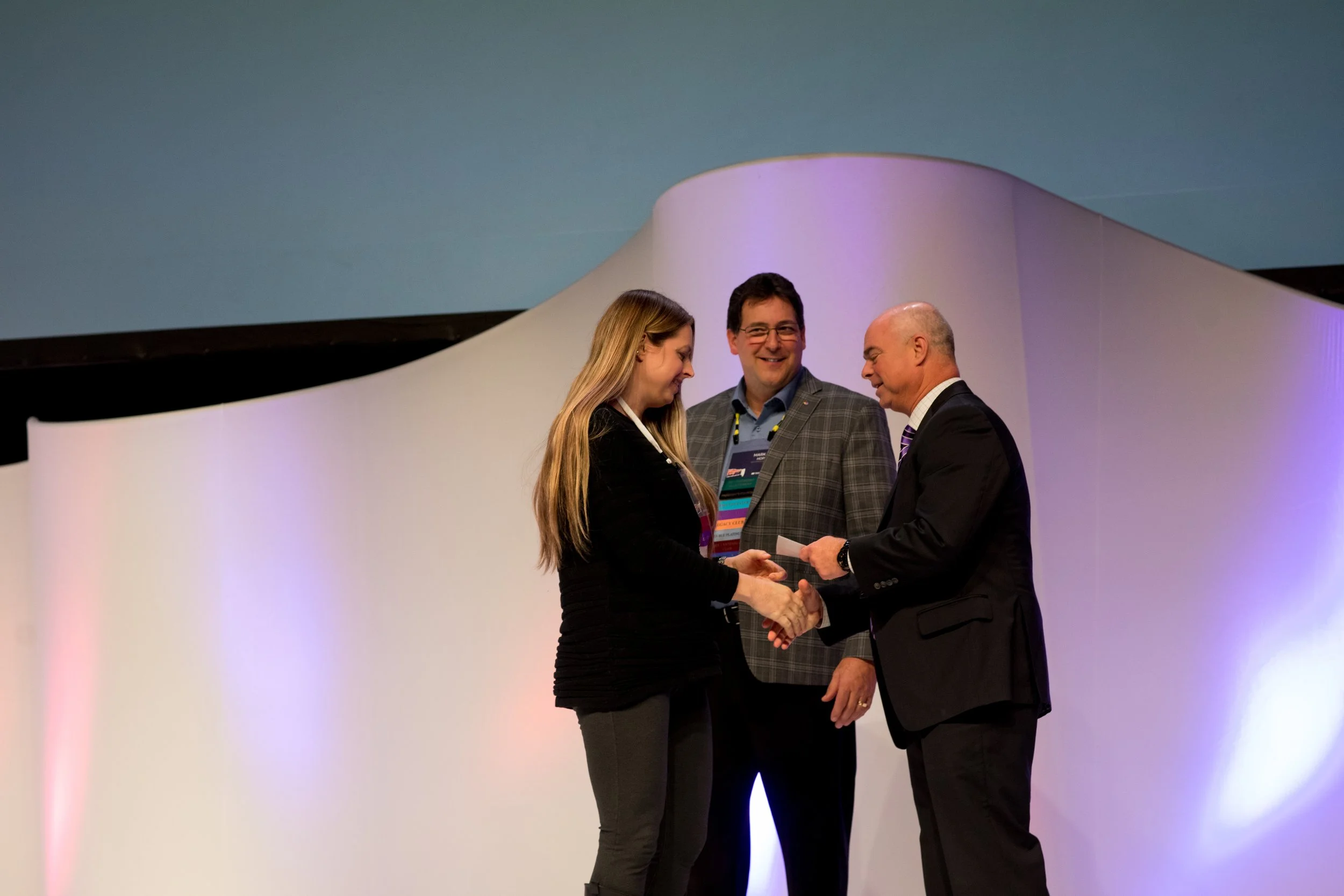 A woman receiving an award or certificate from an older man on stage, with a man standing behind them.