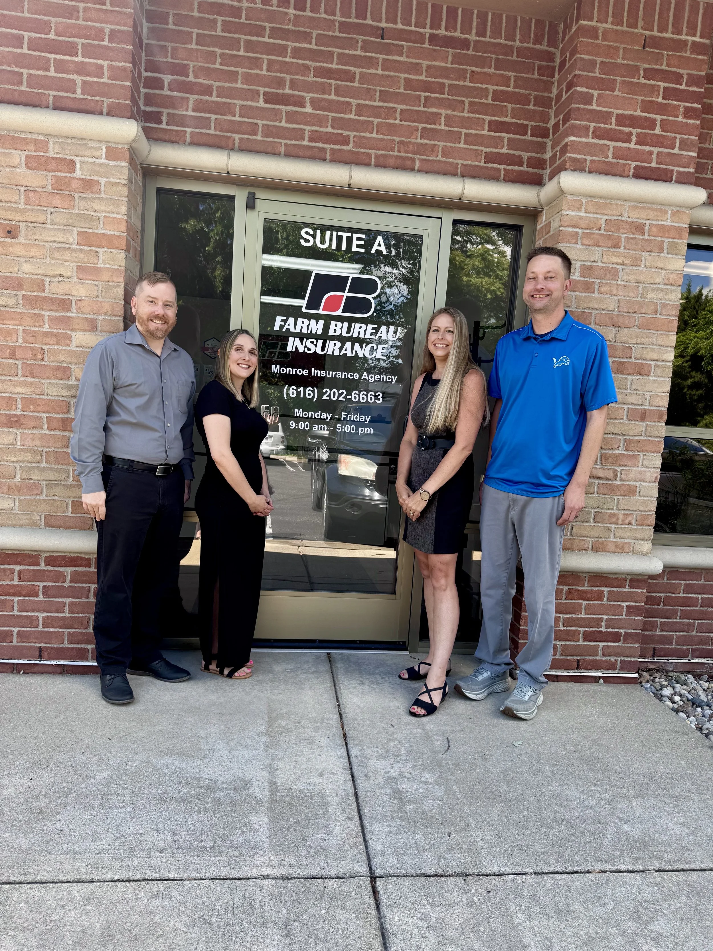Group of five people standing outside in front of a brick building, smiling at the camera. The building has a glass door with the sign for FARM BUREAU INSURANCE, and three of the people are women and two are men, dressed in business casual attire.