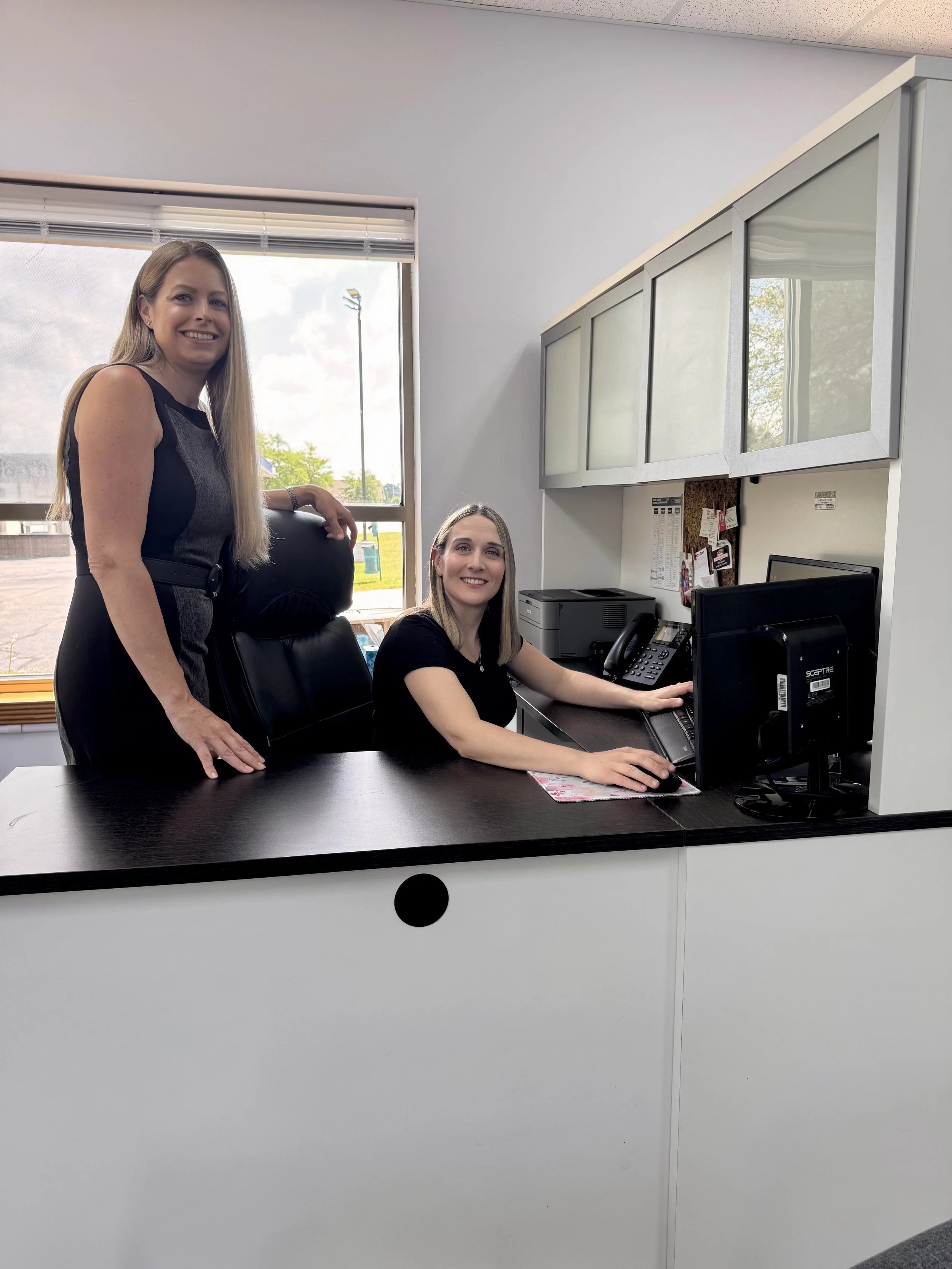 Two women in an office, one standing and one sitting at a desk, with computer monitors, a phone, and office supplies visible.