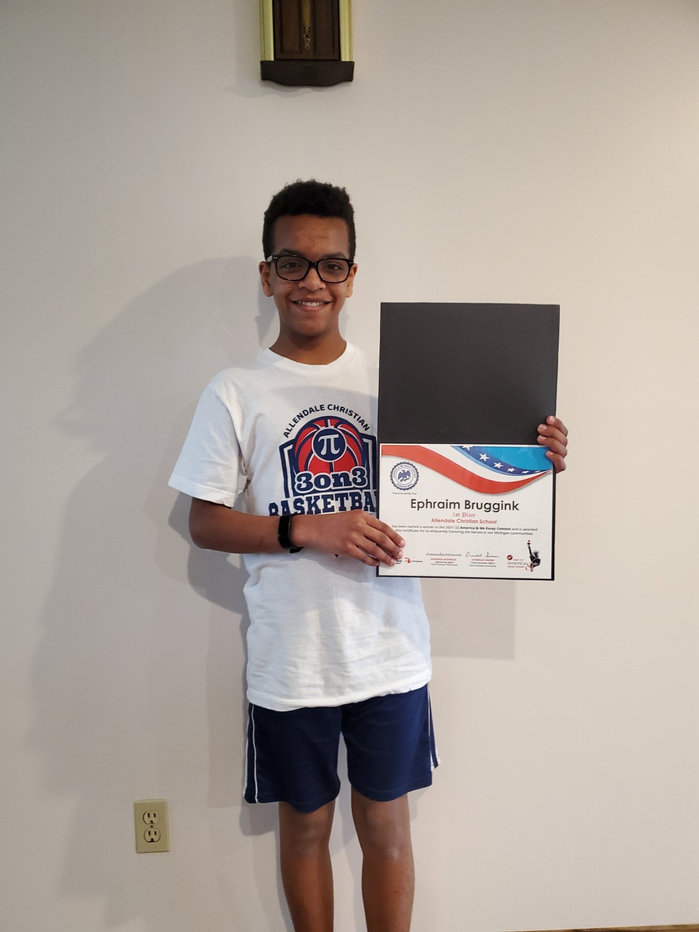 Young man with glasses and short curly hair smiling, holding a certificate in front of a plain wall.