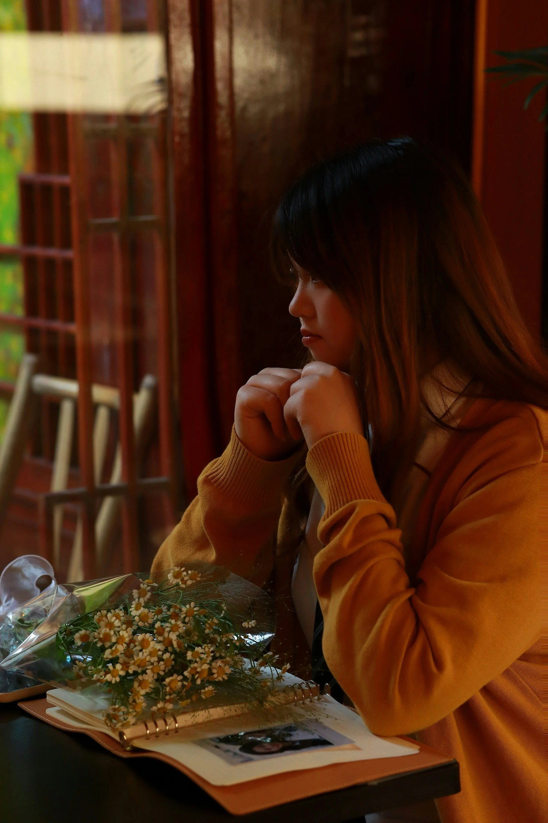 Mulher de cabelos longos e escuros, usando suéter amarelo, olhando para uma floricultura com buquê de margaridas, em um ambiente com iluminação quente e móveis de madeira.