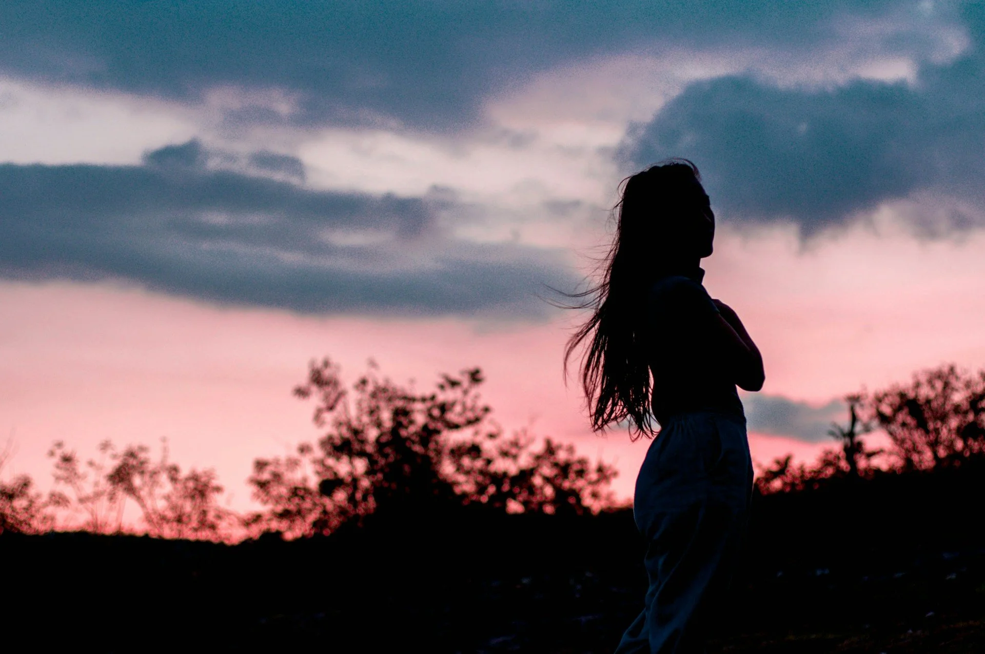 silhueta de uma mulher com cabelo longo ao pôr do sol com céu de nuvens e árvores no fundo