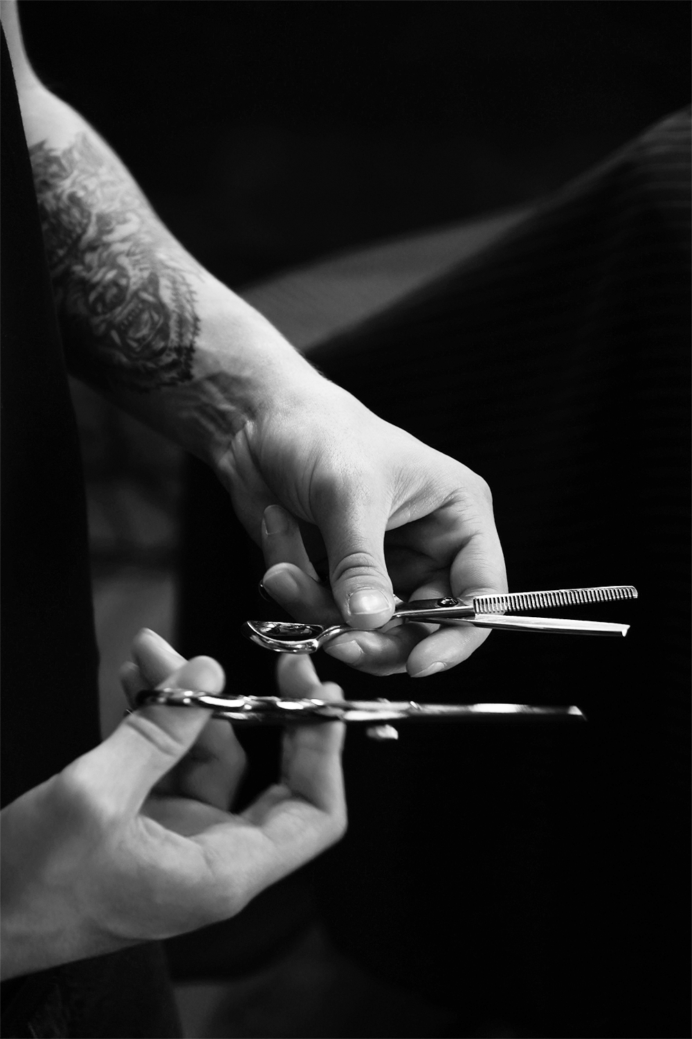 Manos sosteniendo tijeras y pinzas de barbería o para corte de cabello, cerca de un tatuaje en el antebrazo en un ambiente oscuro y nublado.