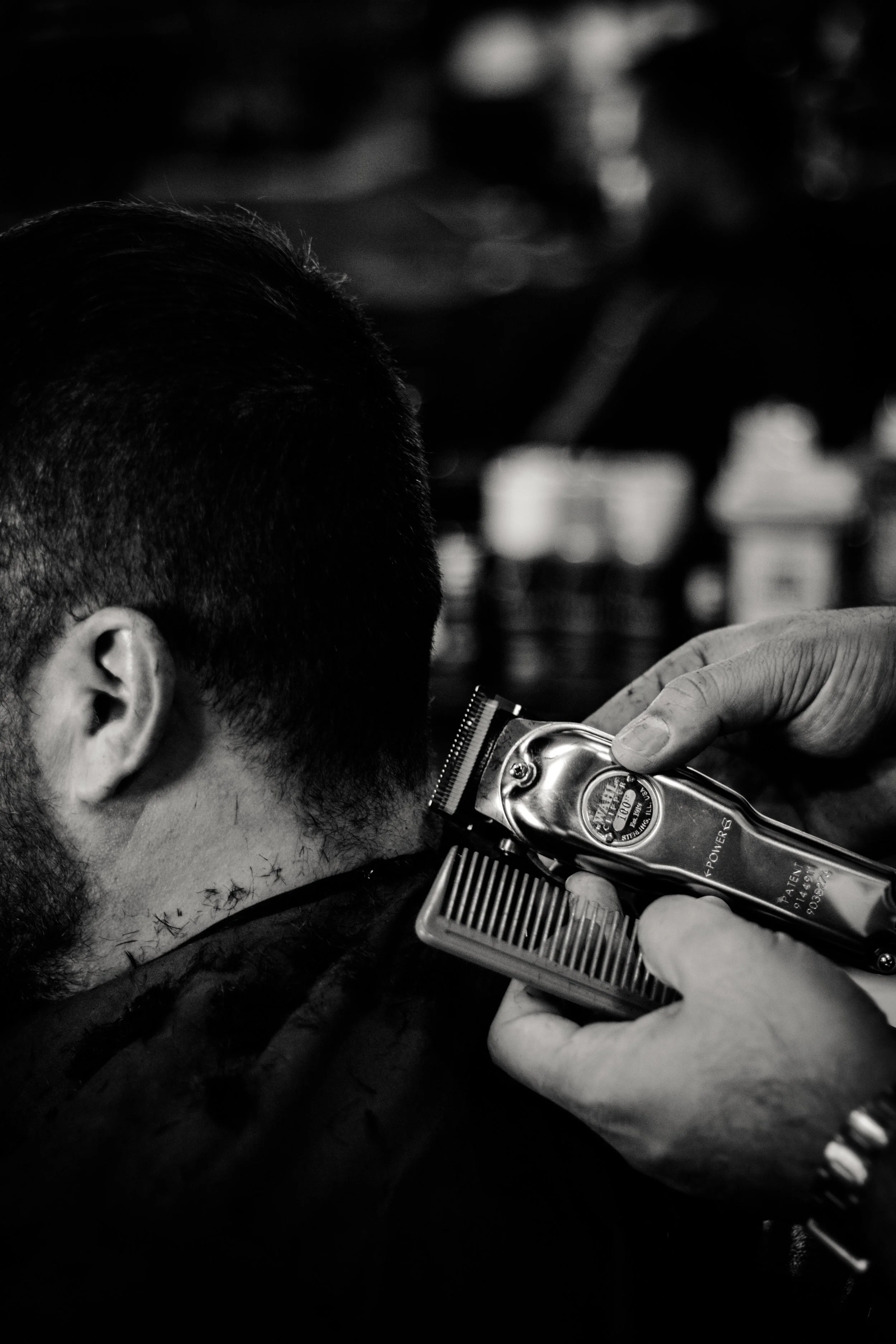 Un barbero cortando el cabello de un cliente con una máquina eléctrica, en un entorno de barbería. La imagen es en blanco y negro y muestra solo la parte superior de la cabeza y la mano del barbero sosteniendo la máquina.