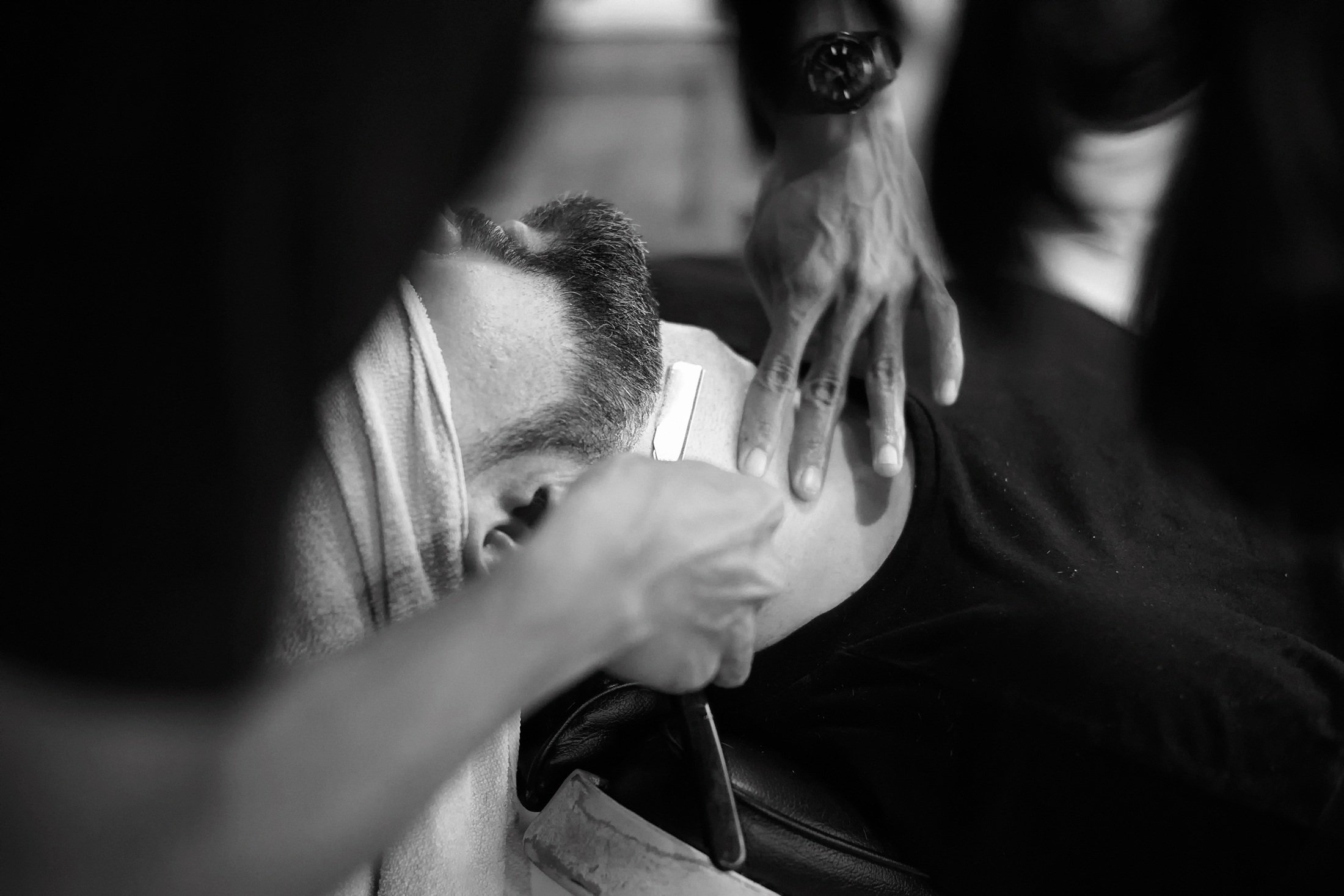 Una persona con barba y cabello corto recostada en un asiento de automóvil con ojos cerrados, mientras otra persona con piel clara y cabello oscuro le toca la cabeza y la cara, en un entorno en blanco y negro.