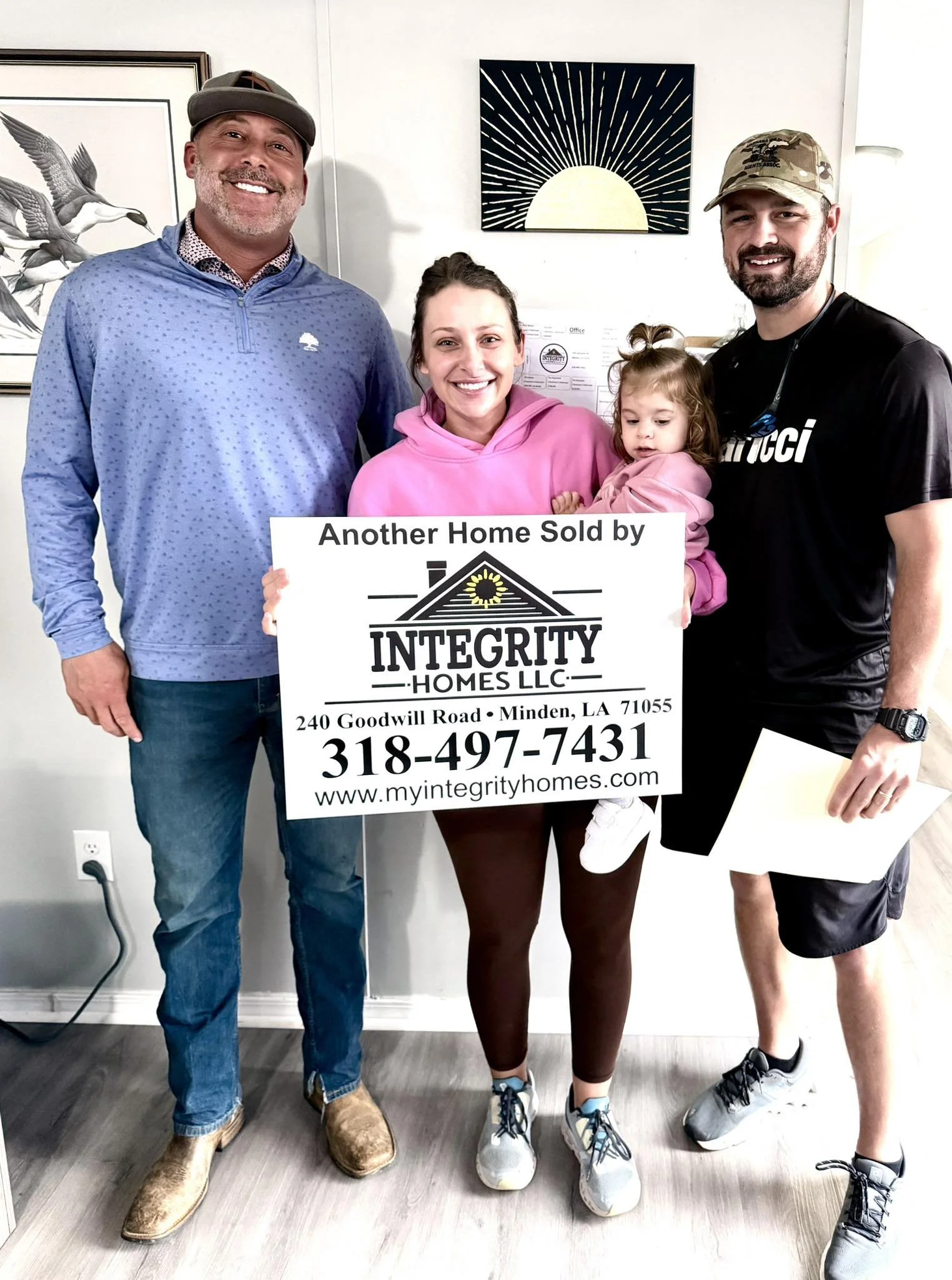 A group of four people standing indoors, holding a sign that reads 'Another Home Sold by Integrity Homes LLC', with a woman and a little girl in the middle and two men on either side; they are smiling.