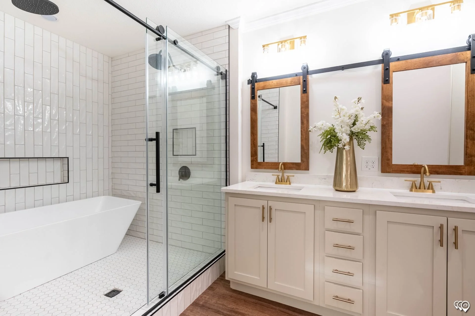 A modern bathroom featuring a walk-in glass shower with white subway tile walls, a white bathtub, and a double vanity with two mirrors and gold fixtures. There is a decorative vase with white flowers on the vanity. The space has wood accents and appears bright with natural light.