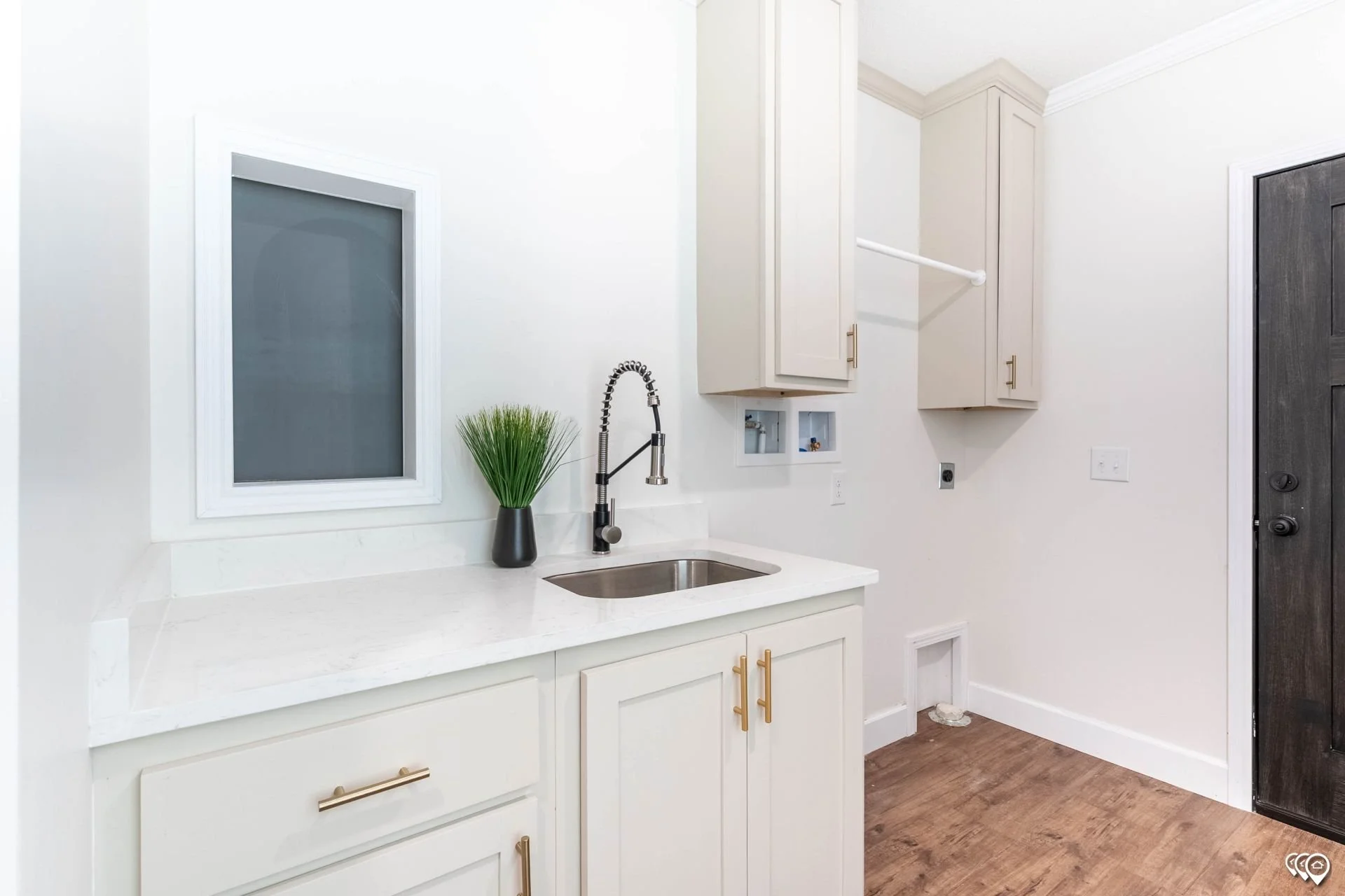 A modern white kitchen with closed cabinets, a black door, a small window, and a steel sink with a black faucet. There is a potted plant on the white countertop.