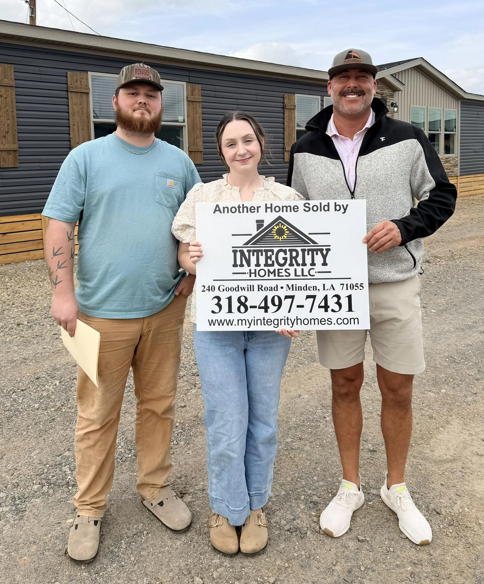 Three people standing outdoors in front of a house. They are posing for a photo while holding a sign that reads, 'Another Home Sold by Integrity Homes LLC.' The person on the right is wearing a black and gray jacket, beige shorts, and white sneakers. The woman in the middle is wearing a beige blouse and blue jeans. The man on the left is dressed in a light blue t-shirt, tan pants, and beige shoes. They are smiling or have neutral expressions.