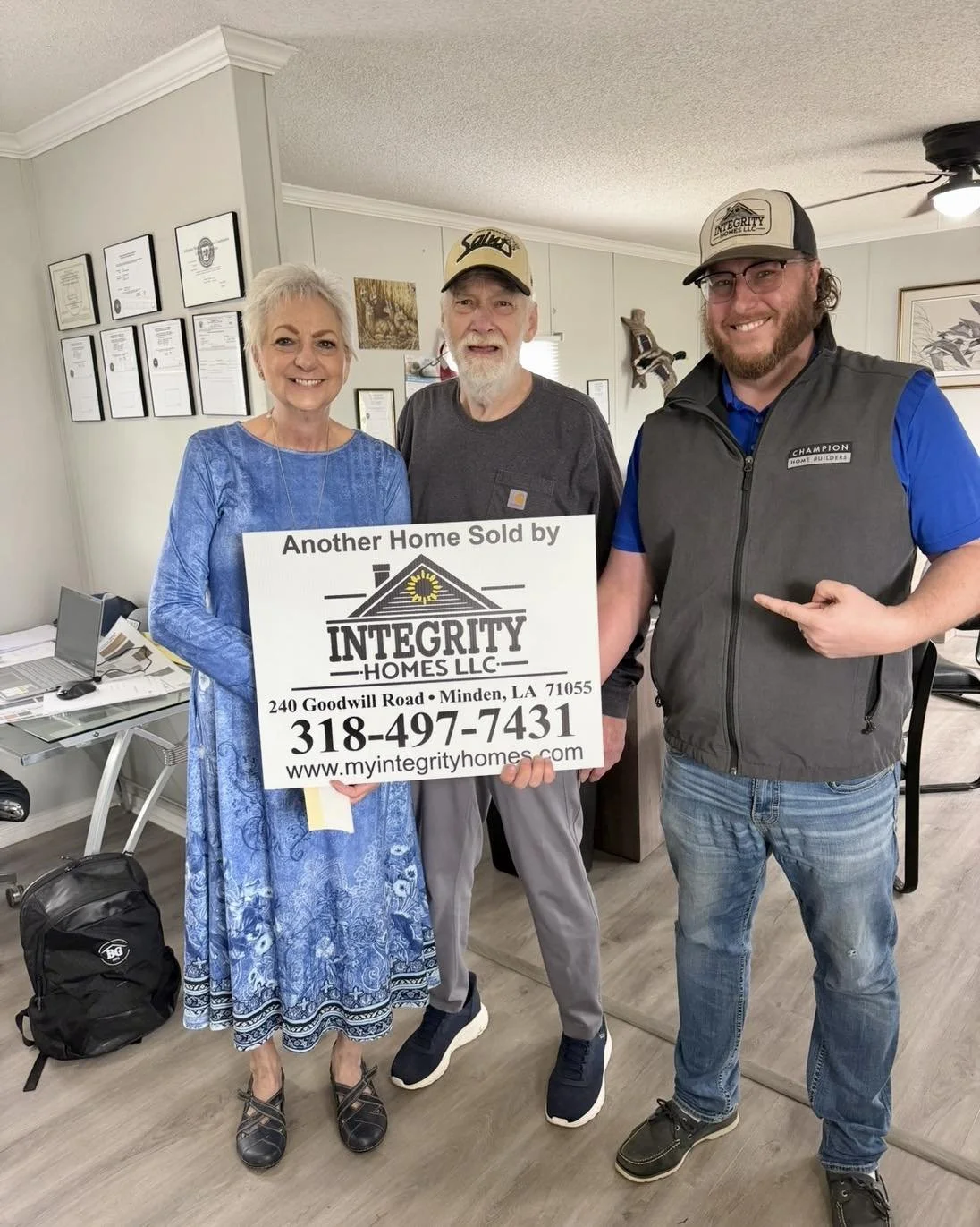 Three people standing in an office, holding a sign that reads 'Another Home Sold by Integrity Homes LLC'. The woman on the left is wearing a blue dress, and the man in the middle is wearing a gray shirt and baseball cap. The man on the right is weari