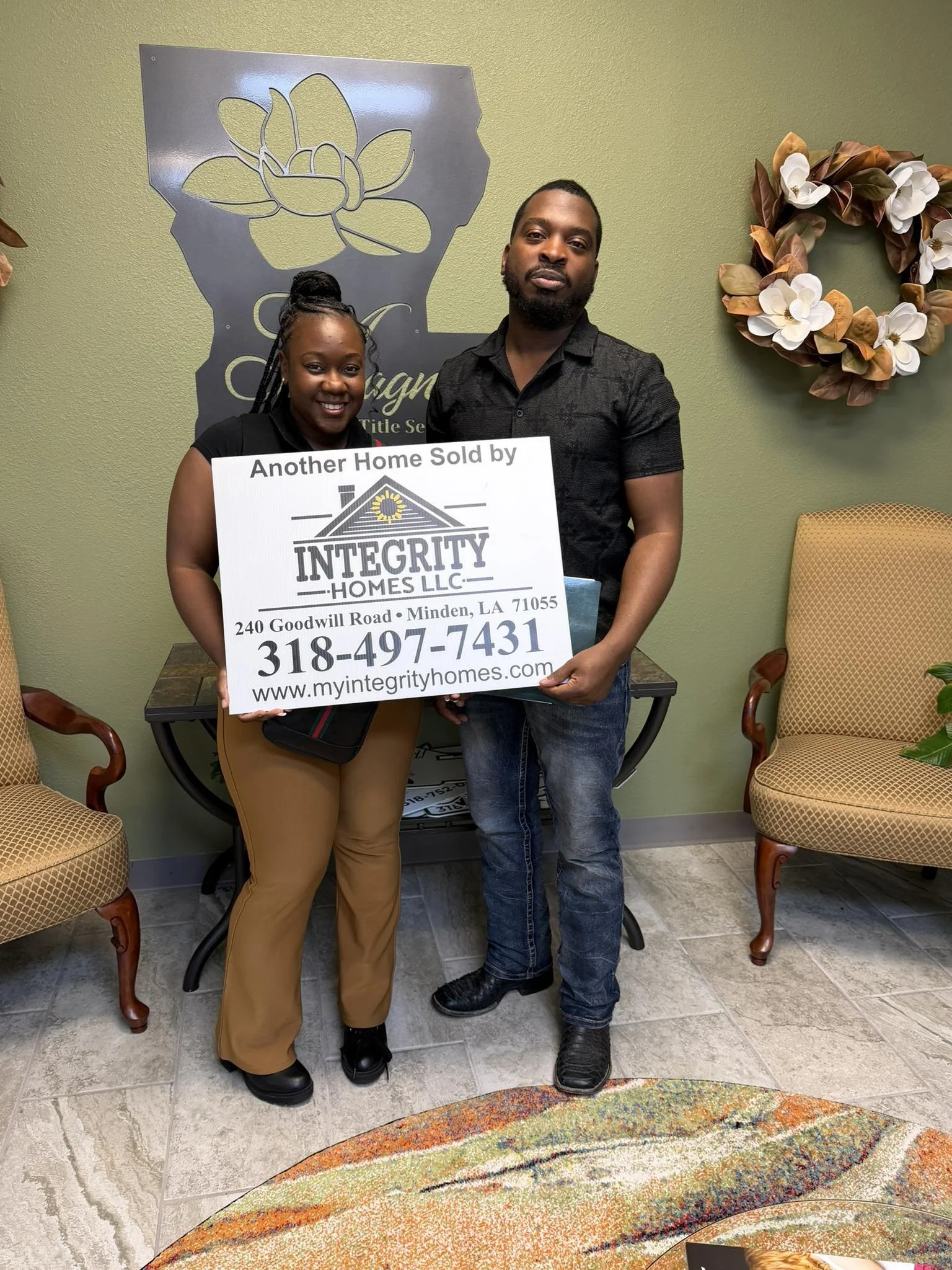 A man and a woman standing inside a room holding a sign for Integrity Homes LLC. The woman is wearing tan pants and black shoes, and the man is dressed in jeans and black shoes. There are chairs and a decorative wreath on the wall behind them.