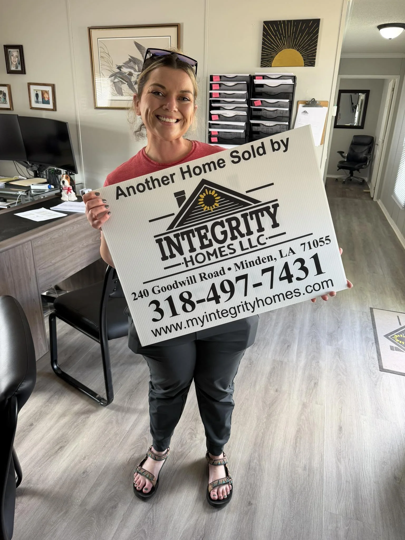 Woman smiling and holding a sign for Integrity Homes LLC in an office with artwork and a desk.