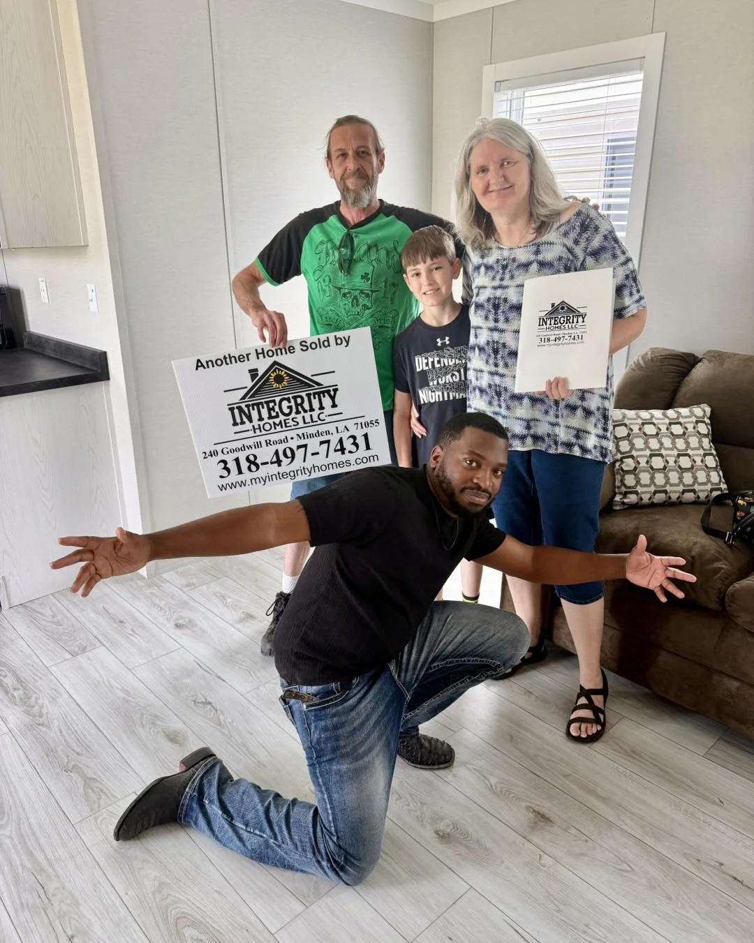 Group of four people inside a home, celebrating the sale of a house. One person is kneeling on the floor with arms wide open, two are standing holding signs that read 'Another Home Sold by Integrity Homes LLC,' and a young boy is standing between the