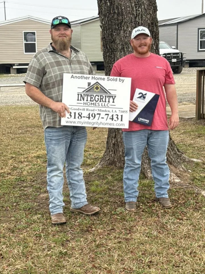 Two men standing outdoors in front of a large tree. One man is holding a sign for Integrity Homes LLC, advertising a home sale in Minden, Louisiana. The other man holds a folder or pamphlet. Both are smiling and dressed casually. In the background, t