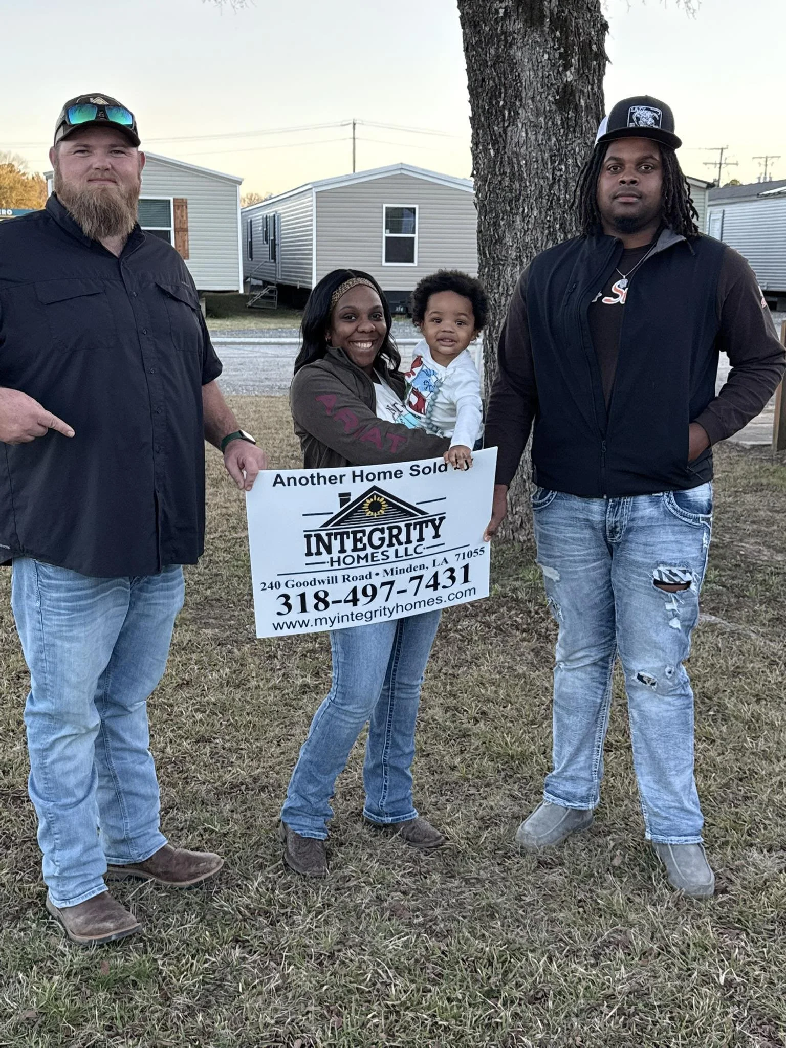 Four people, three men and a woman holding a small child, standing outdoors next to a tree, holding a sign that says 'Another Home Sold' and has the logo and contact information for Integrity Homes. They are dressed casually and appear happy.